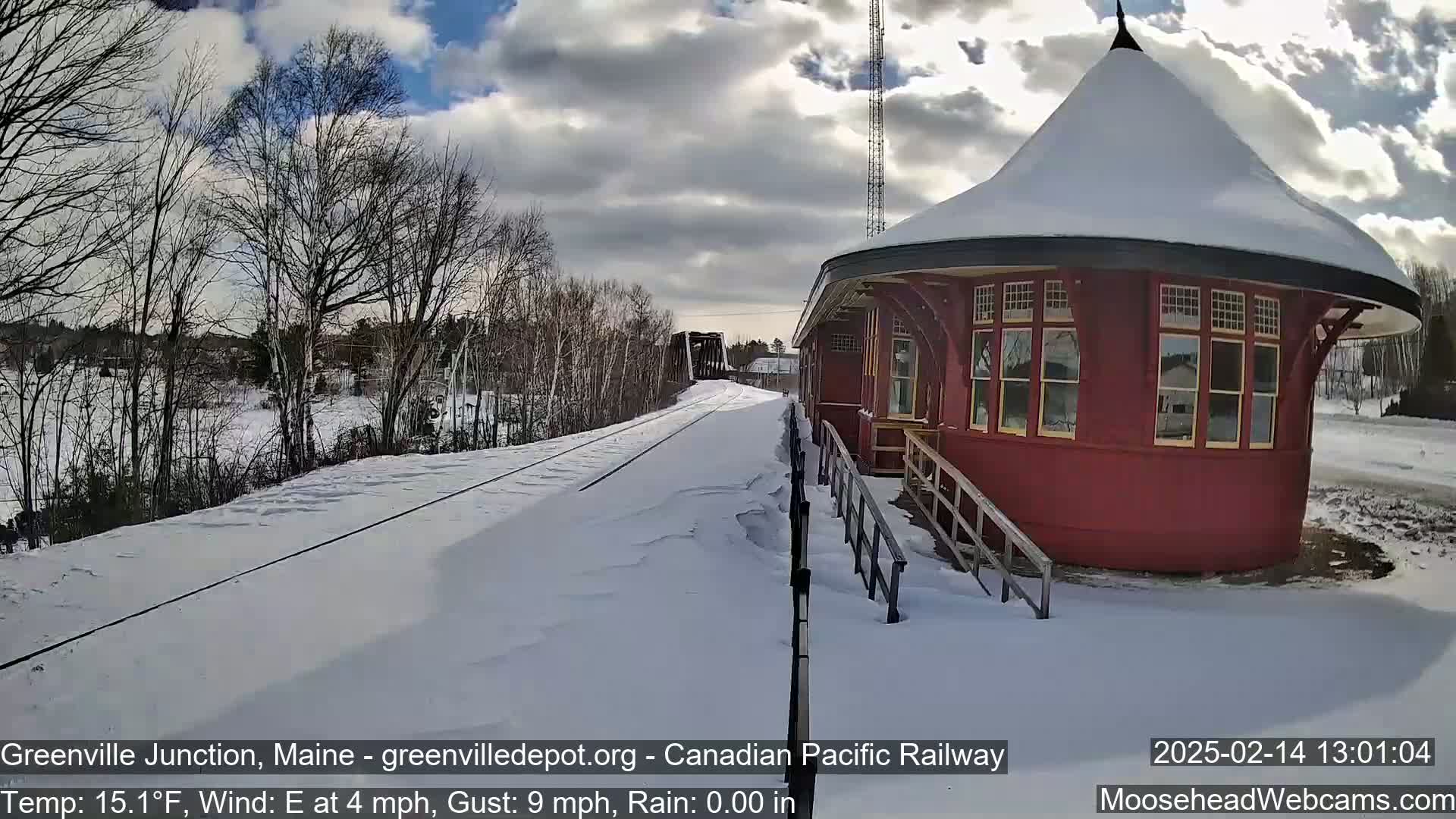 A red train station sits beside snow-covered train tracks under a partly cloudy sky, with a train visible in the distance.