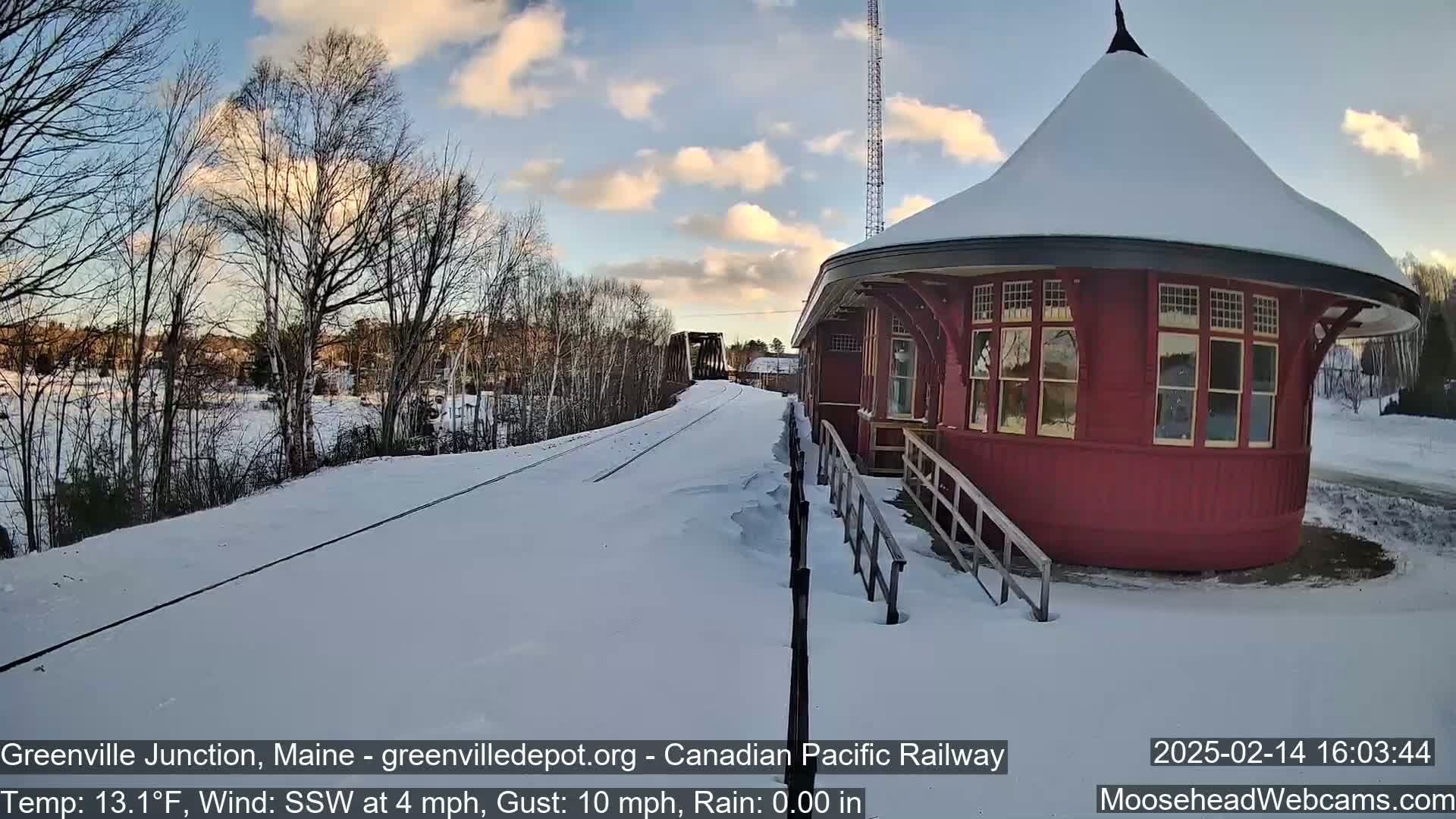 A snow-covered train platform with a red, round depot and a view of train tracks extending toward a bridge under a partly cloudy sky.