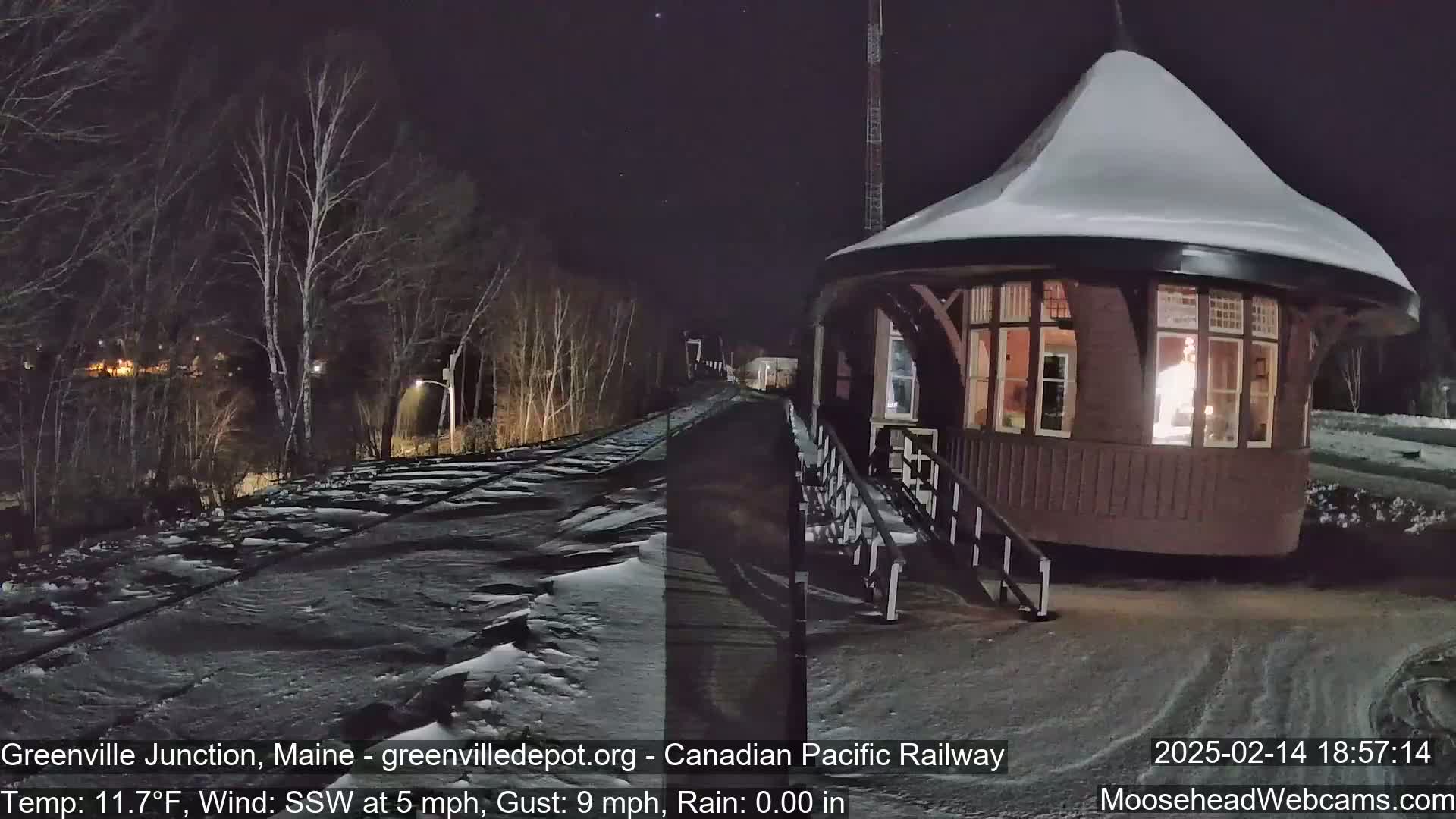 A snow-covered, round train station with a lit interior is situated beside a snow-dusted railway track at night, under a dark, clear sky.