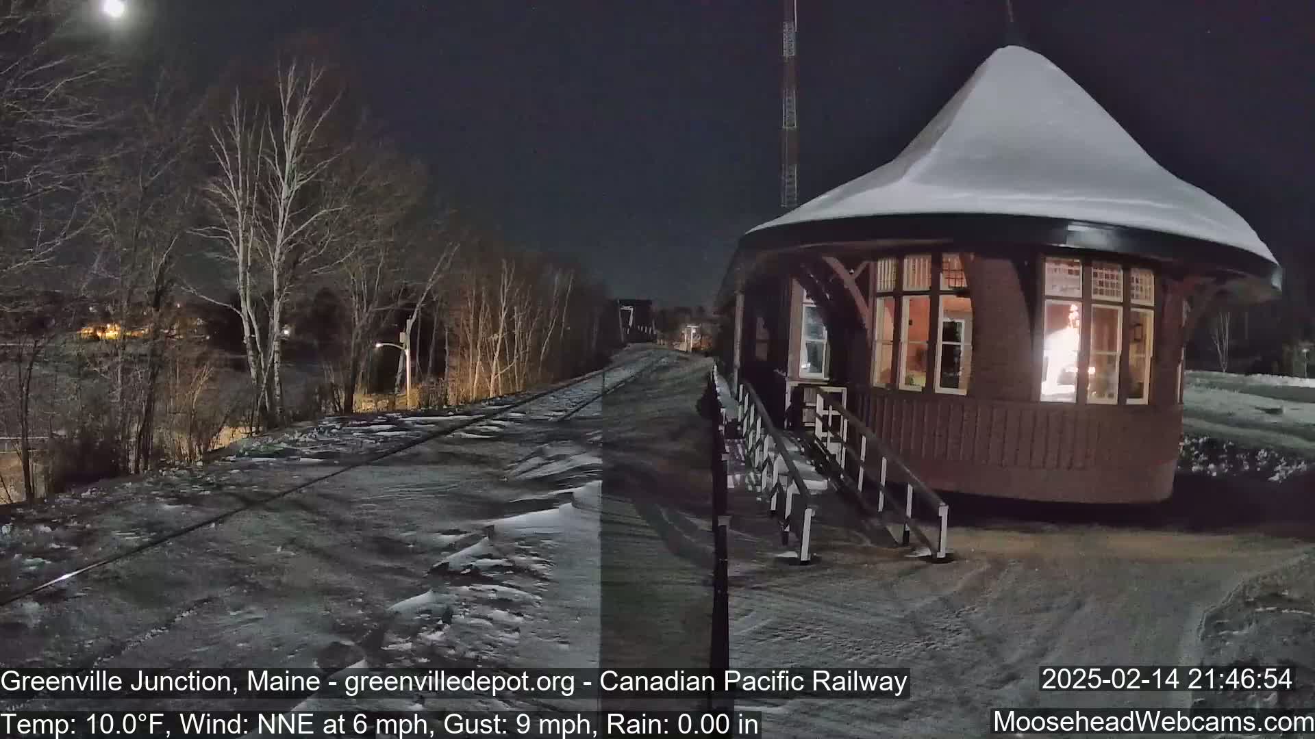 A snow-covered, illuminated train station stands beside a snow-covered railway track under a nighttime sky with a visible moon, in windy, cold conditions.