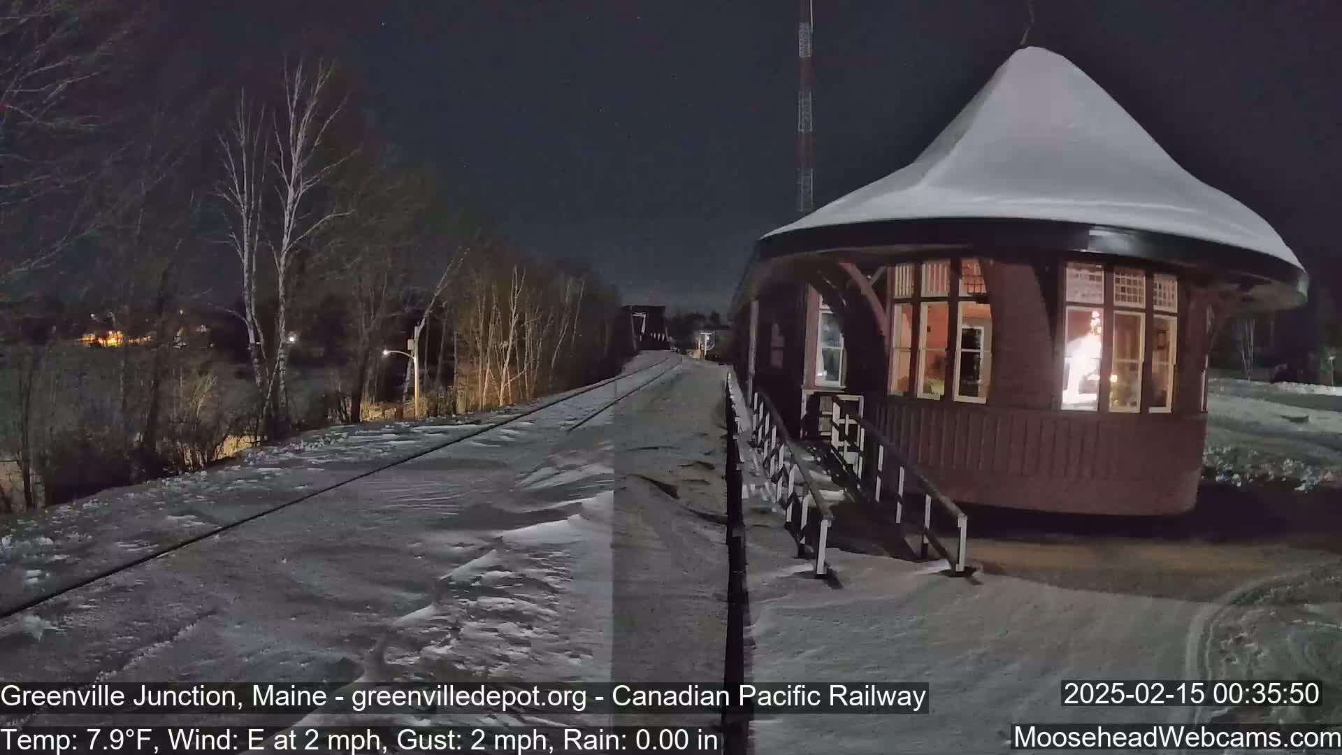 A snow-covered train depot is illuminated at night, next to a snow-covered railway track under a clear, dark sky.