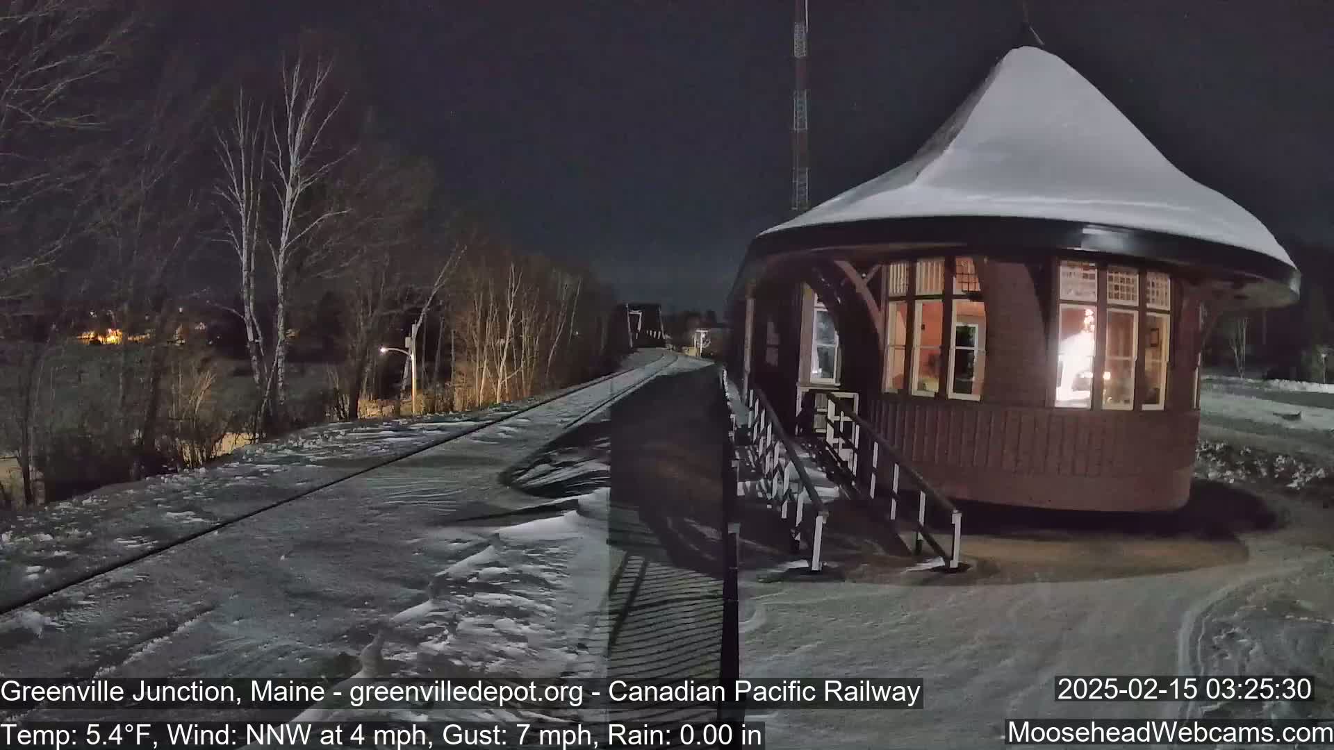 A snow-covered, round train station is illuminated at night next to a snow-covered railway track under a dark sky.