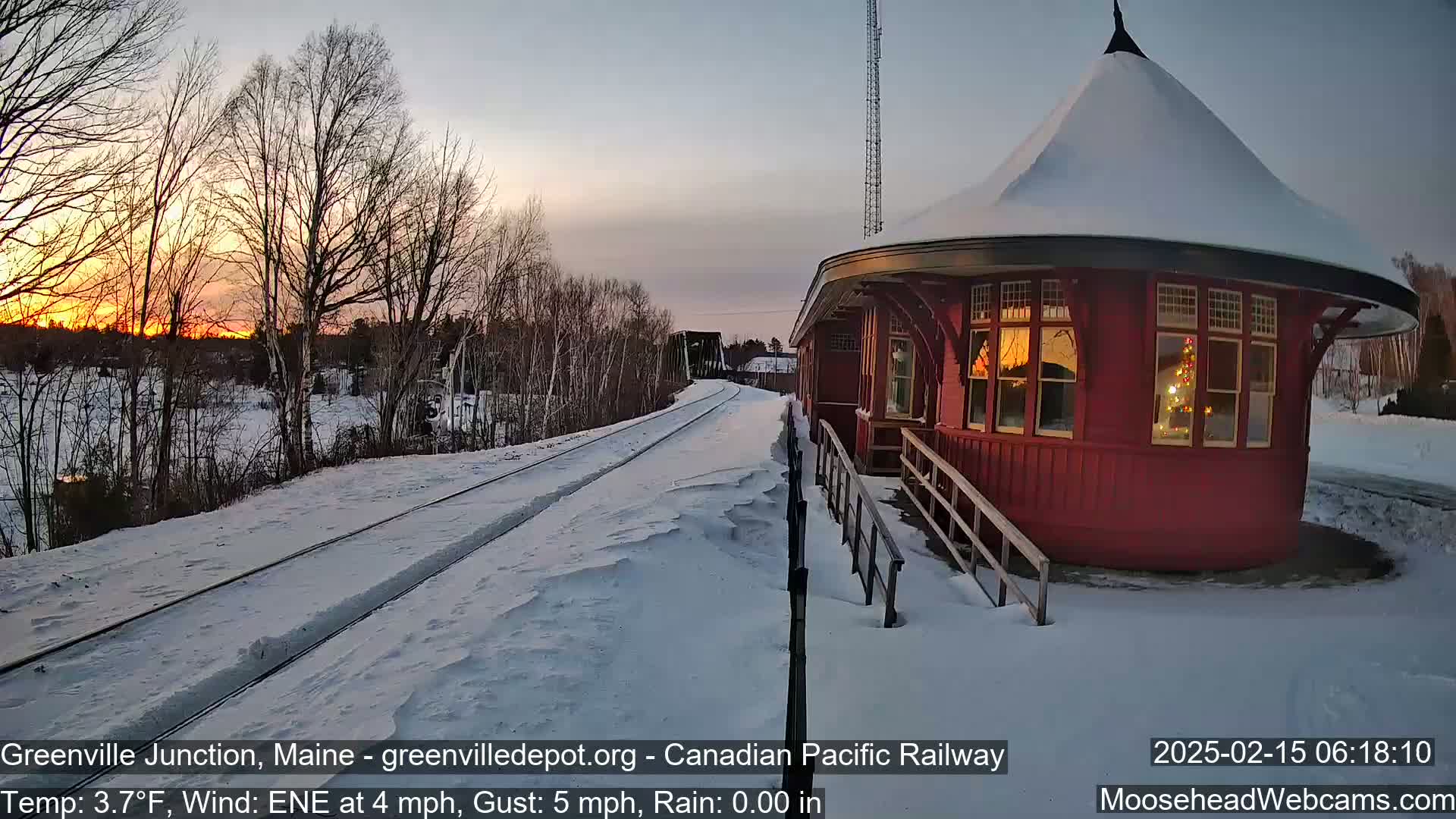 A snow-covered railroad track leads to a red, round train station during a snowy sunset.