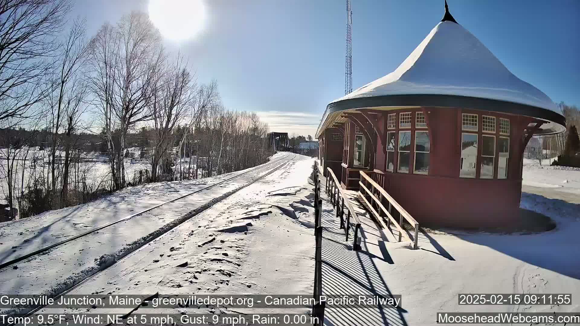 A snow-covered train platform and tracks extend toward a bridge under a bright, sunny sky.