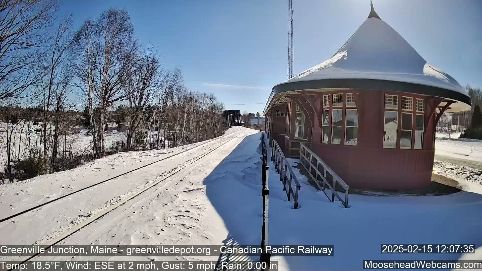 A snow-covered train platform and tracks extend toward a distant bridge under a sunny sky.