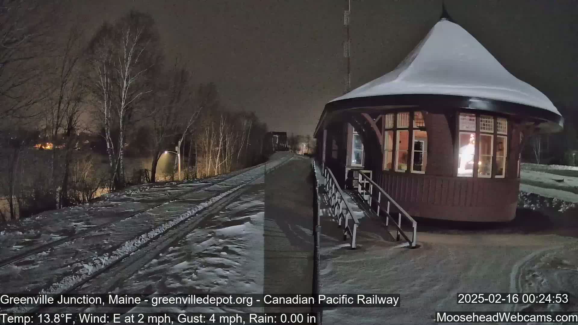 A snow-covered, round train station with a lit interior is situated beside a snow-covered railway track at night.