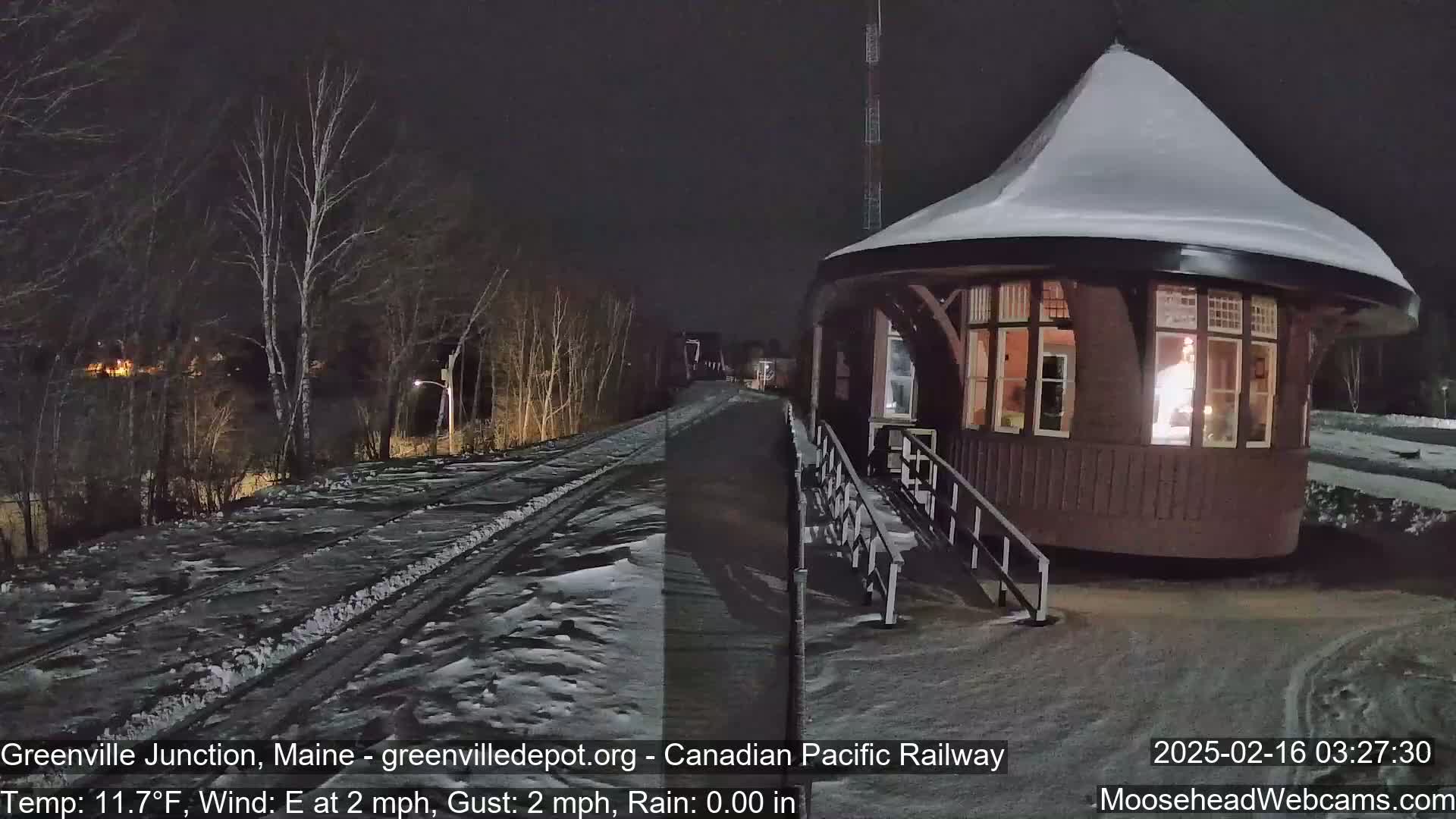 A snow-covered, illuminated train station stands at night beside a snow-dusted railway track, under a dark sky.