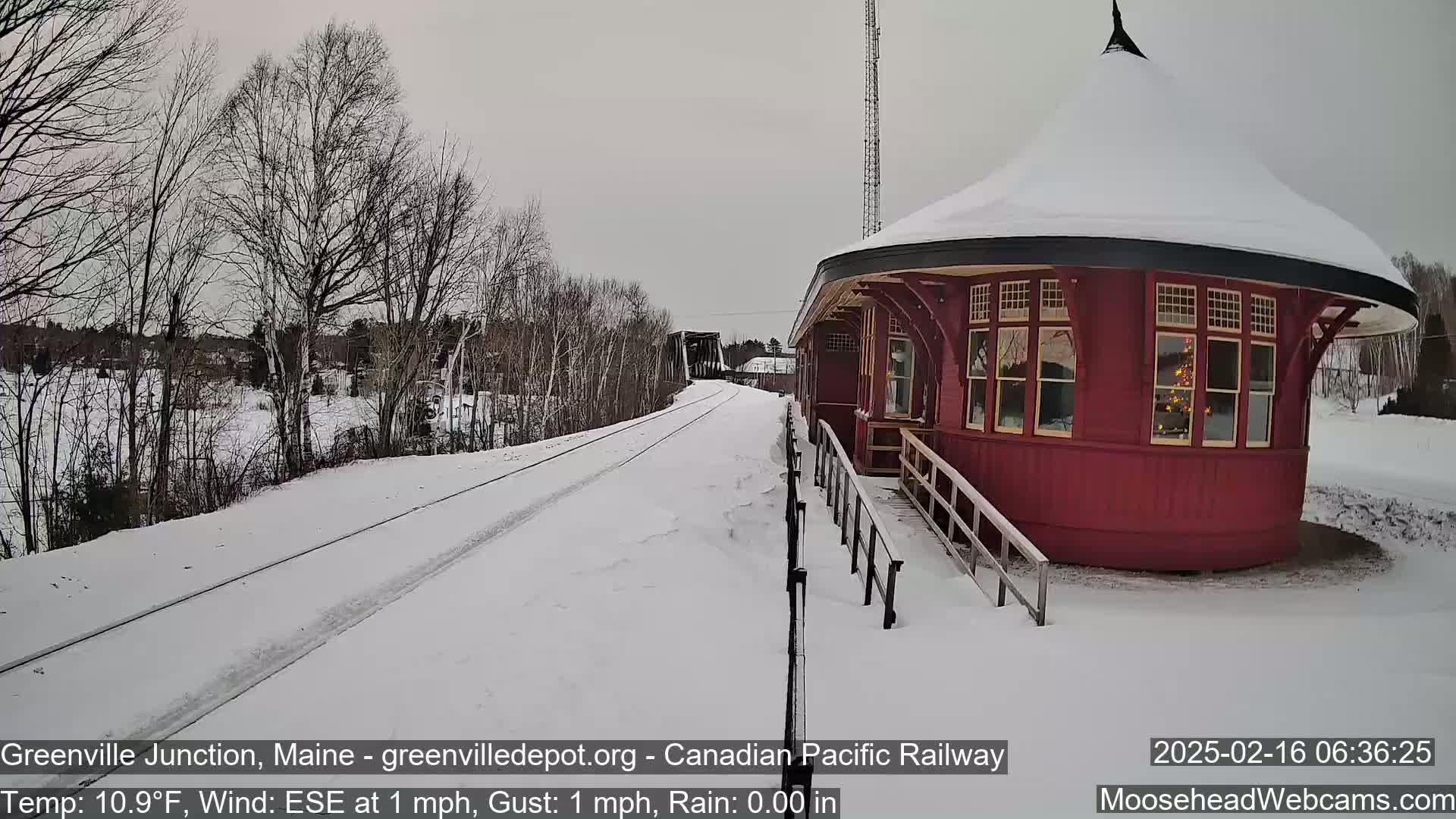 A snow-covered railroad track leads to a round, red train depot under a gray sky.