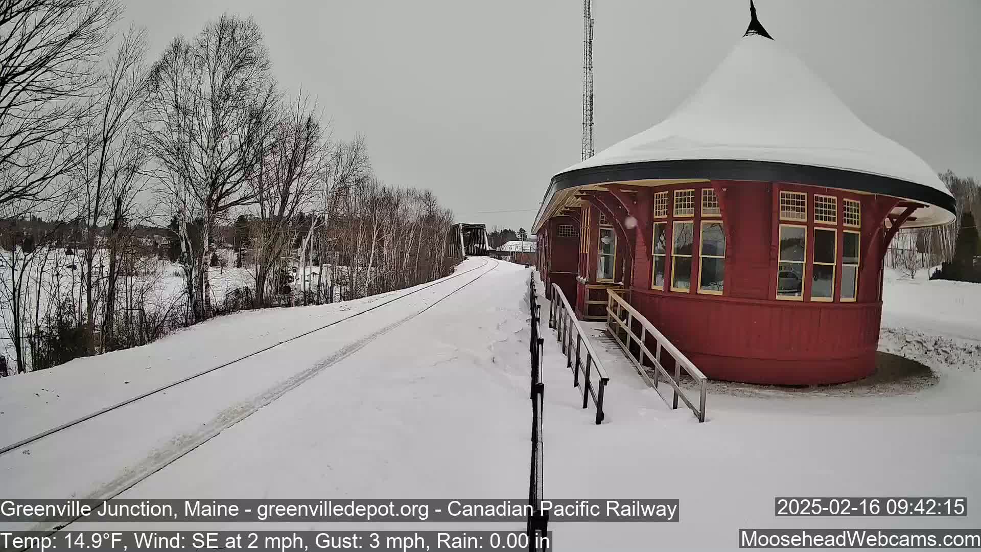 A snow-covered train platform and tracks extend toward a bridge under a gray sky.