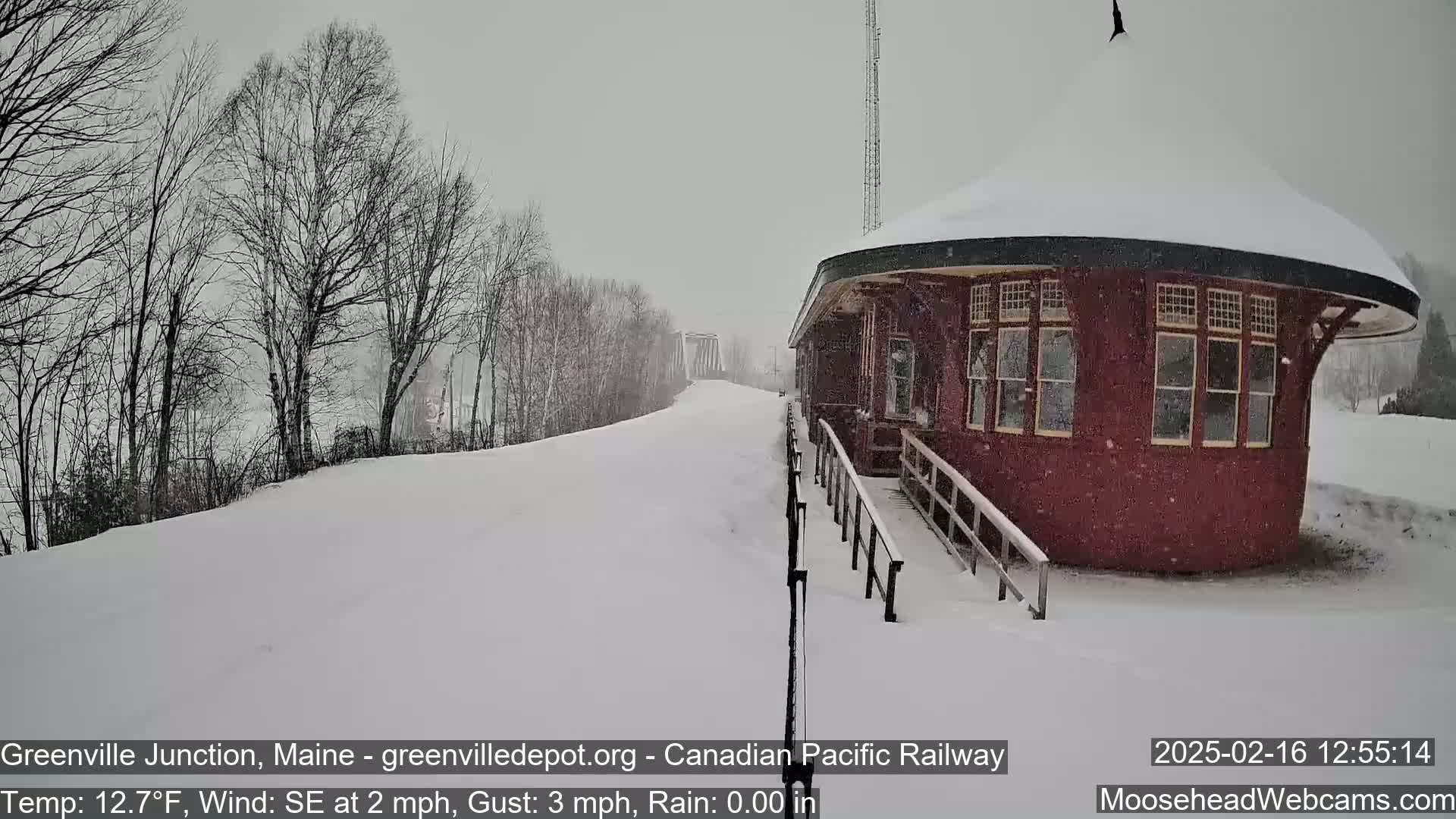 A snow-covered train station sits beside a snow-covered railway track during a light snowfall.