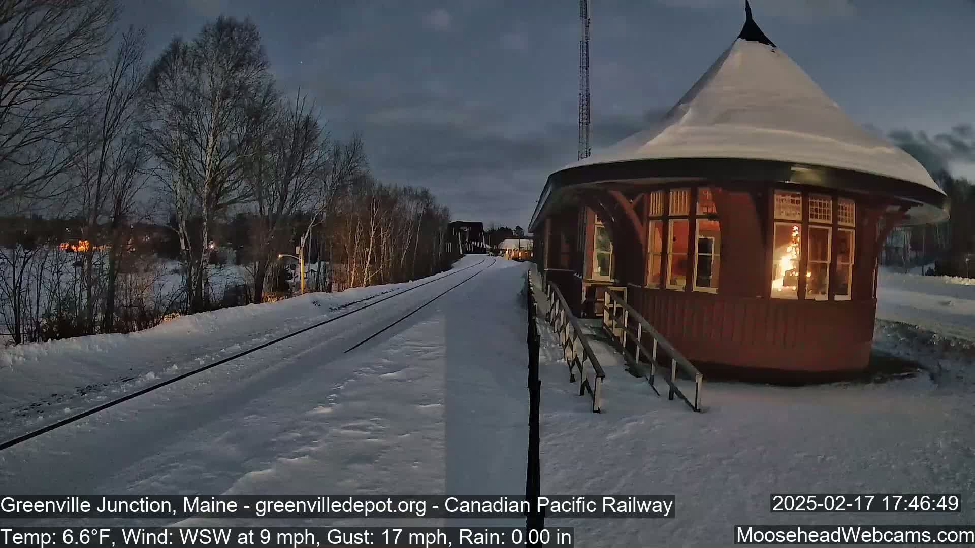 A snow-covered railway track leads to a lit-up, octagonal train station under a twilight sky.