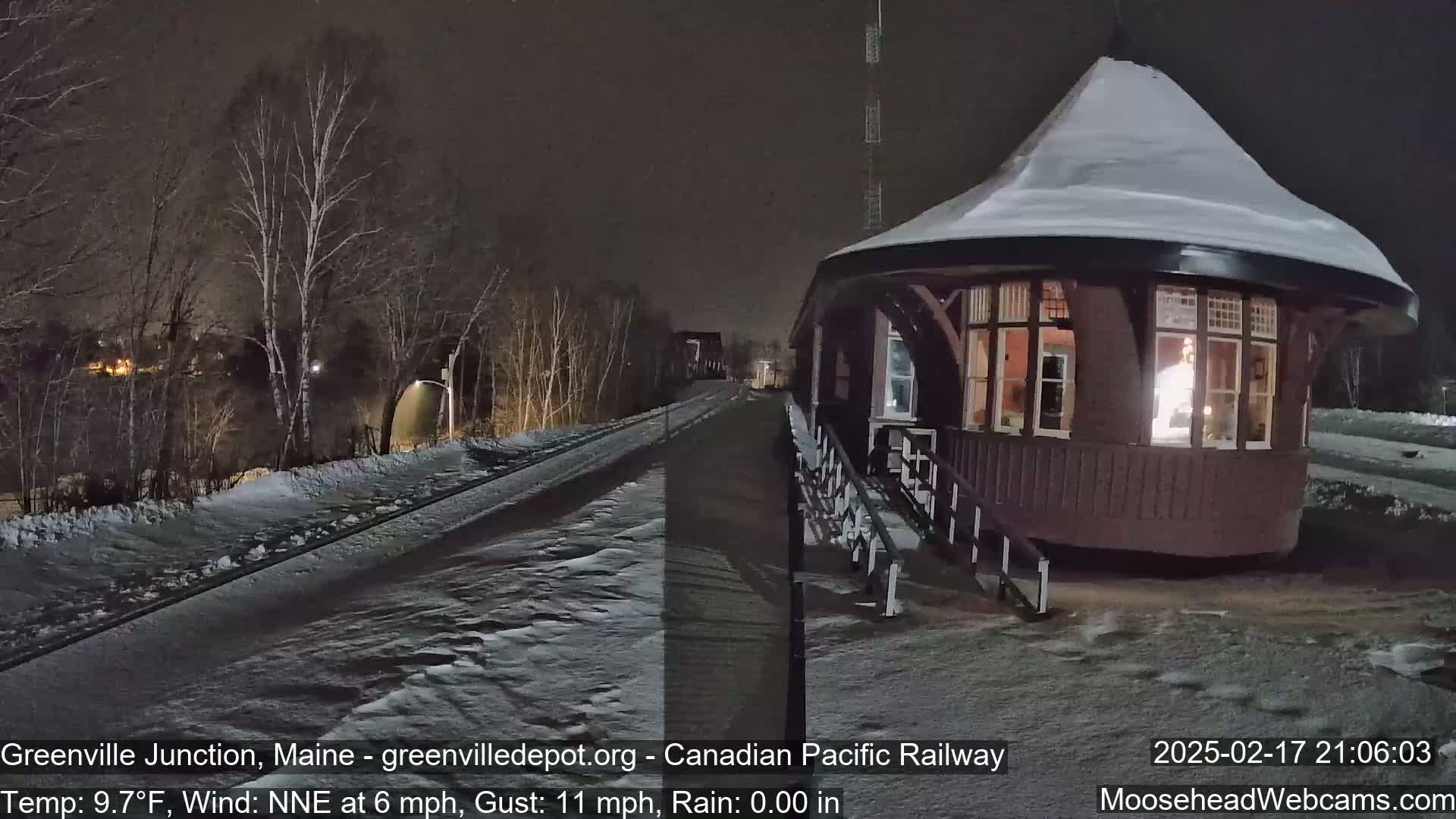A snow-covered, octagonal train depot is illuminated at night next to a snow-covered railway track under a dark sky with a light wind.