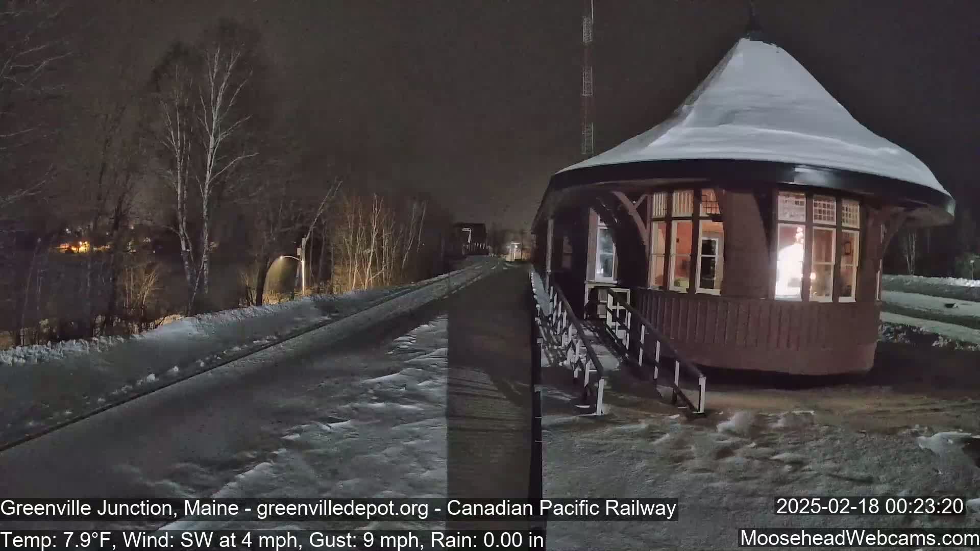 A snow-covered railway station at night, with a train track extending into the background, under a dark sky with a southwest wind.