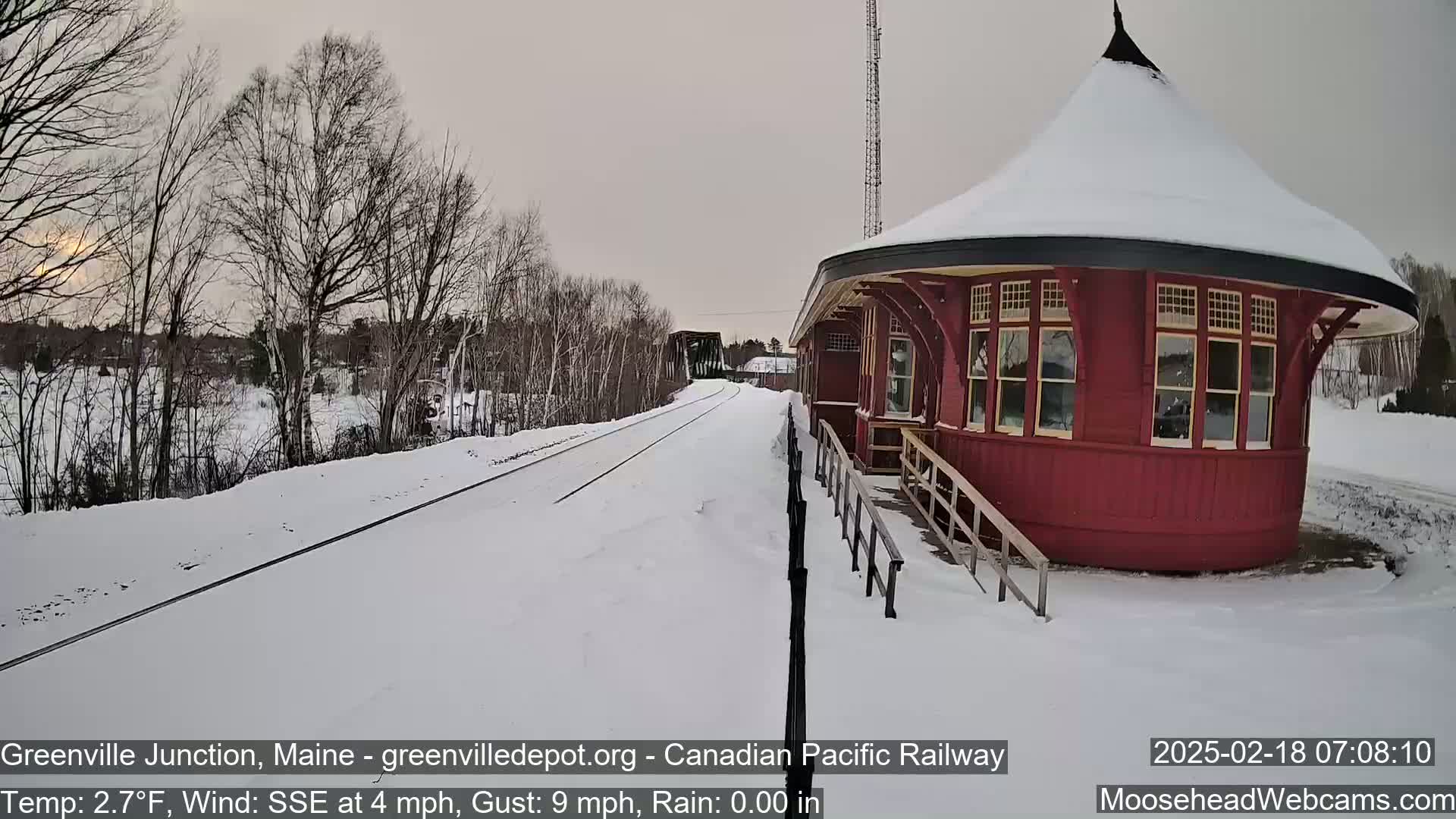A snow-covered train platform with a red circular building is next to a set of train tracks in a winter scene.