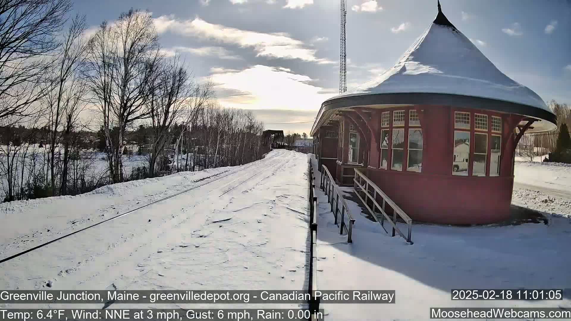 A snow-covered railway platform with a red, round train station is visible under partly sunny skies.