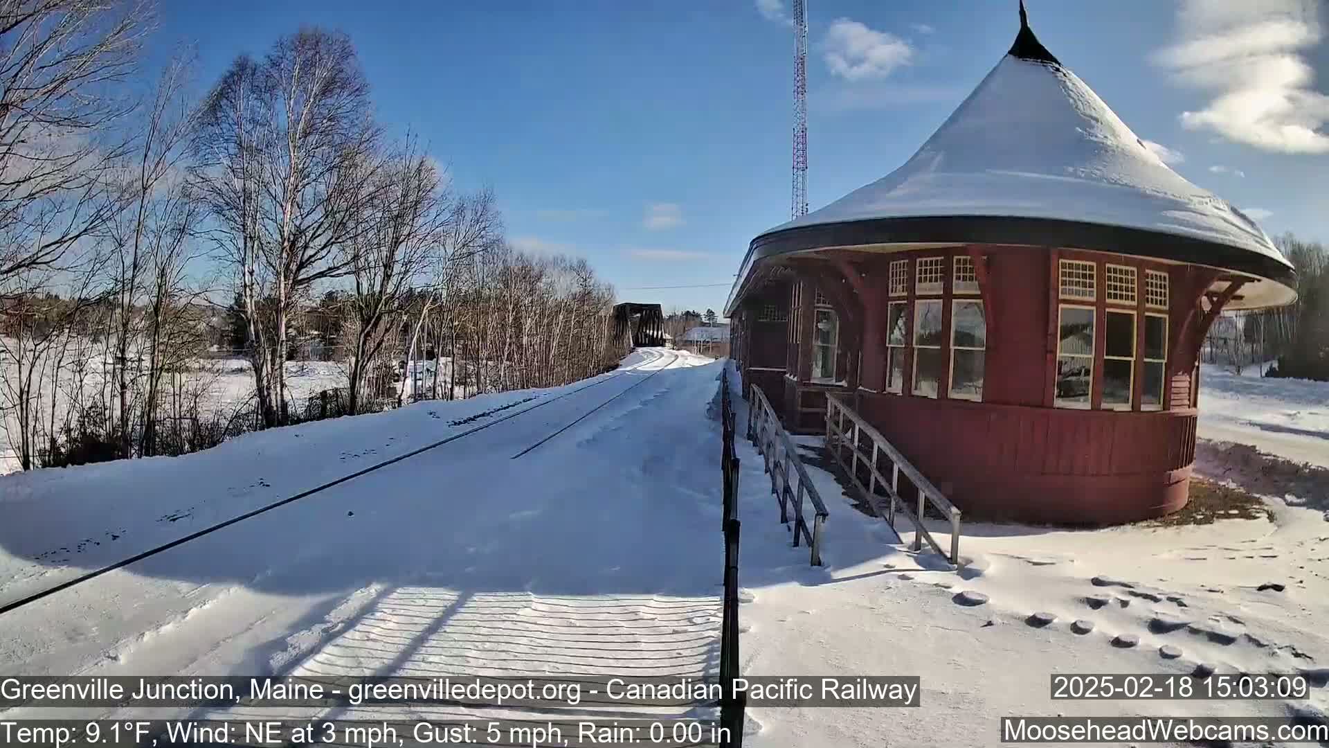 A snow-covered train platform with a round, red train depot is visible under a sunny sky.