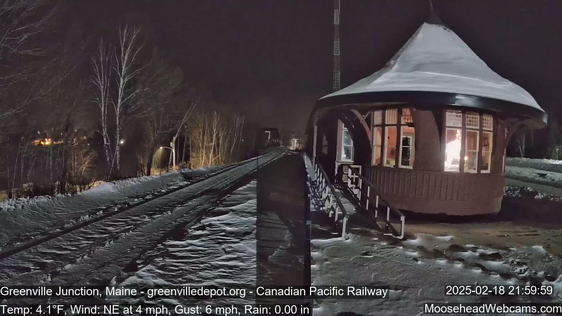 A snow-covered train depot is illuminated at night next to a snow-covered railway track, under a northeast wind.