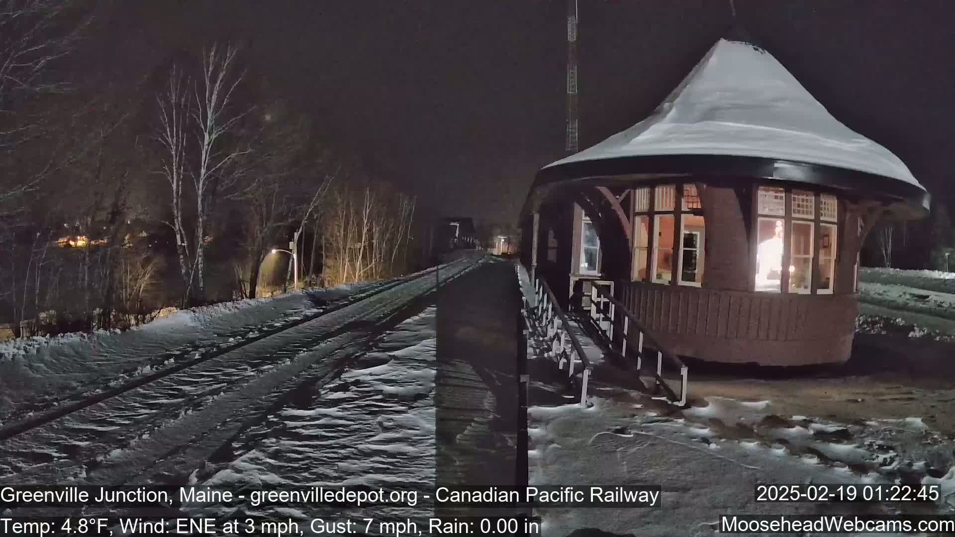 A snow-covered train platform and tracks are visible at night under a dark sky, with bare trees in the background.