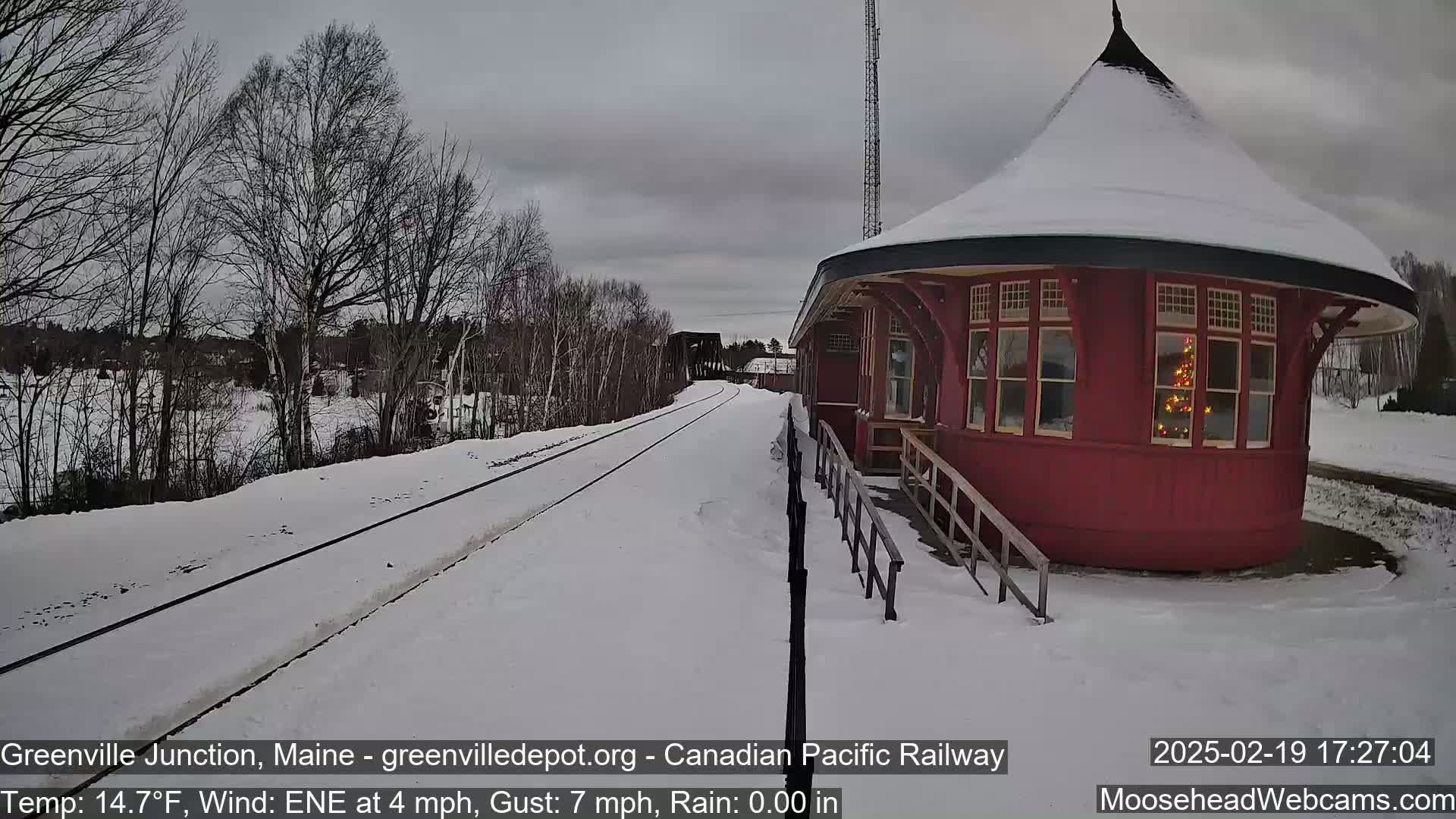 A snow-covered train station with a round, red structure next to snow-covered train tracks under a cloudy sky.
