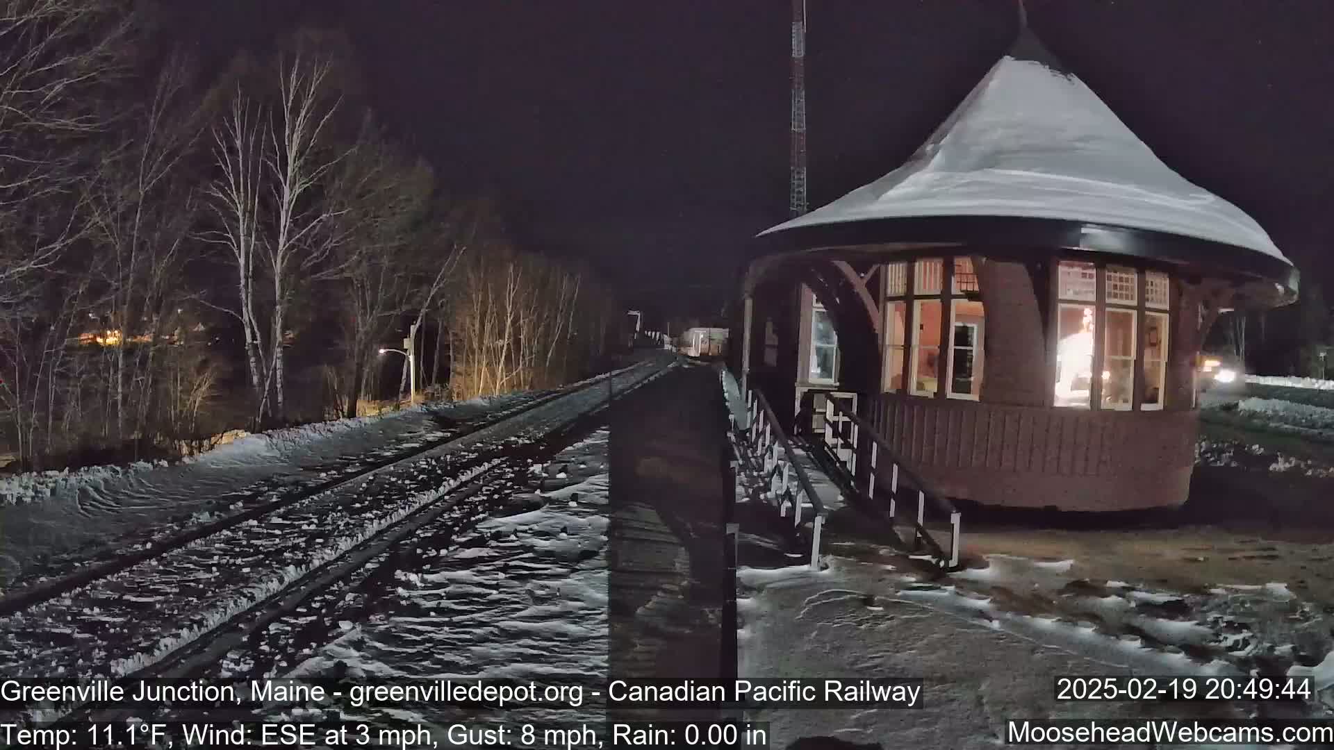 A snow-covered train depot is illuminated at night, with a train track extending into the dark, snowy landscape.