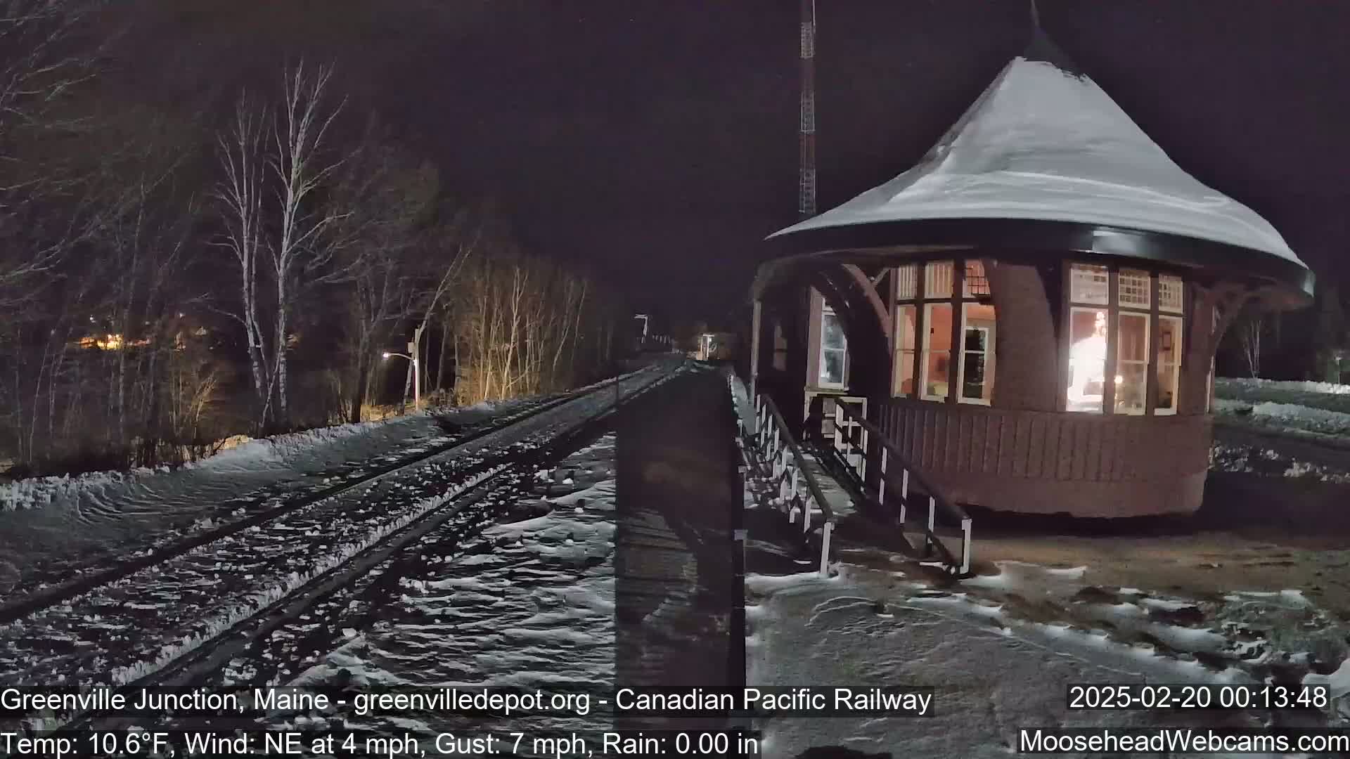 A snow-covered train depot stands beside a snow-dusted railway track at night under a dark sky, with a light northeast wind.