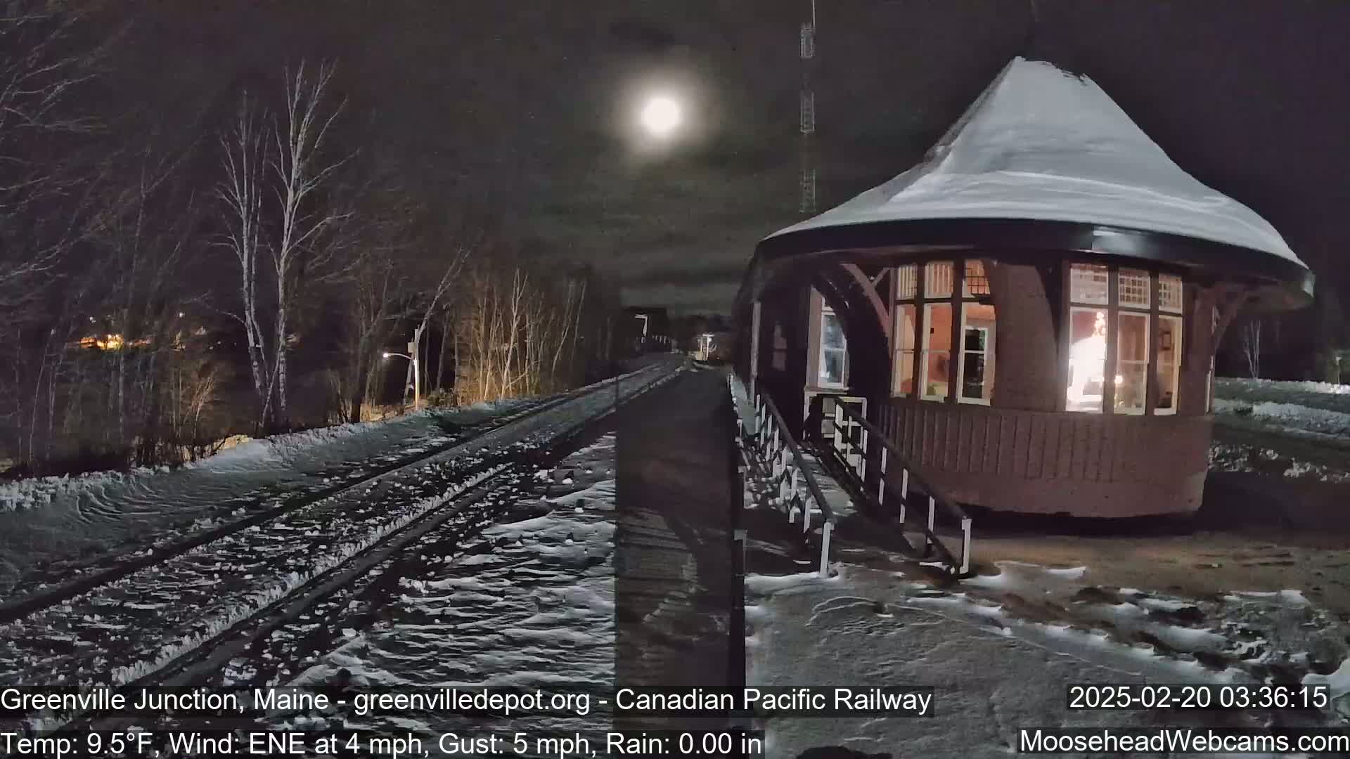 A snow-covered train station with a lit interior is visible at night under a bright moon, with a light snowfall on the tracks nearby.