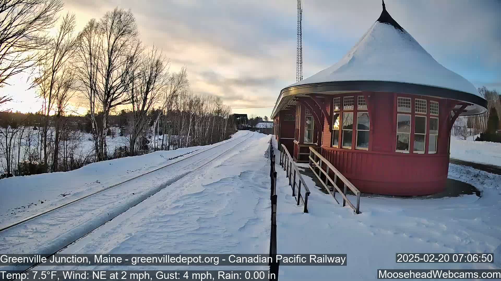 A snow-covered railway platform extends from a round, red train station at sunset, with bare trees and a bridge visible in the background.