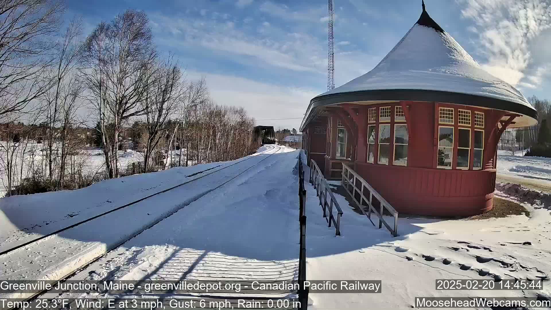 A snow-covered train platform and tracks extend toward a bridge under a sunny sky, with a red, snow-dusted, round train station to the right.