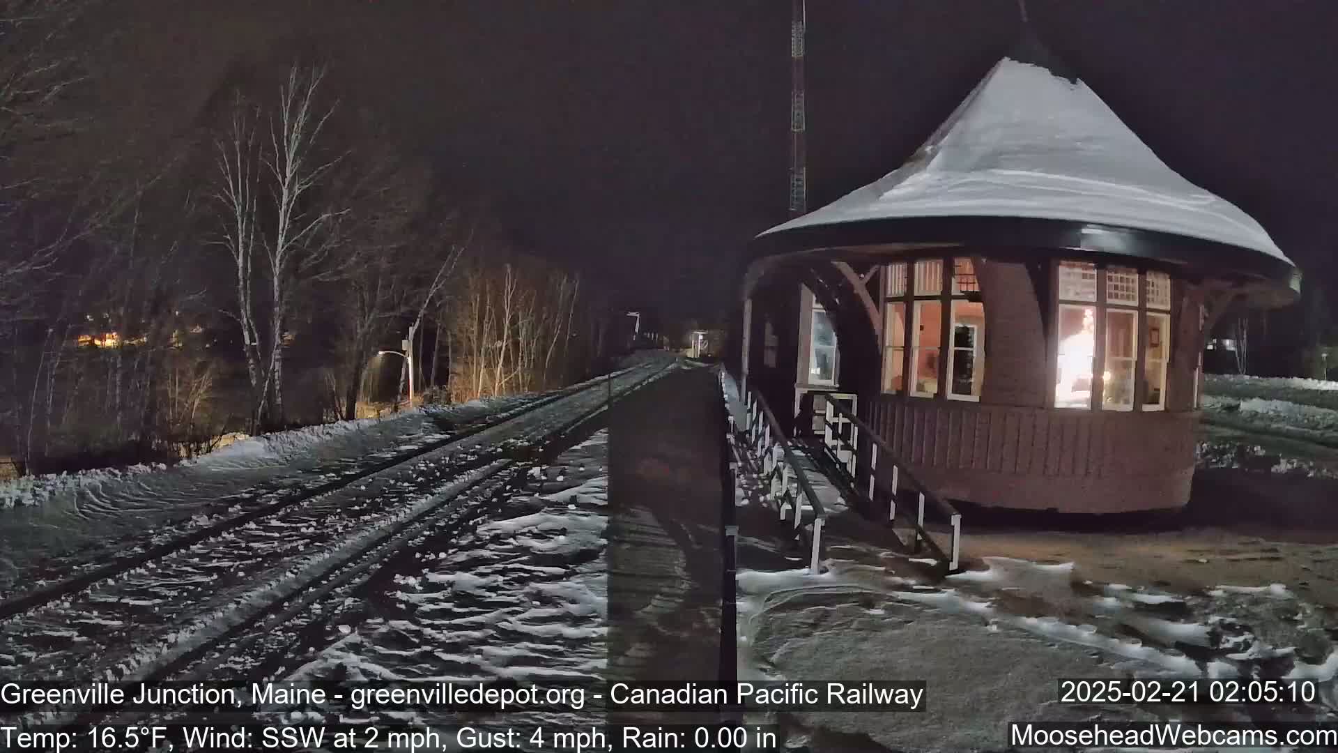 A train passes a snow-covered, round, illuminated building at night in the rain.