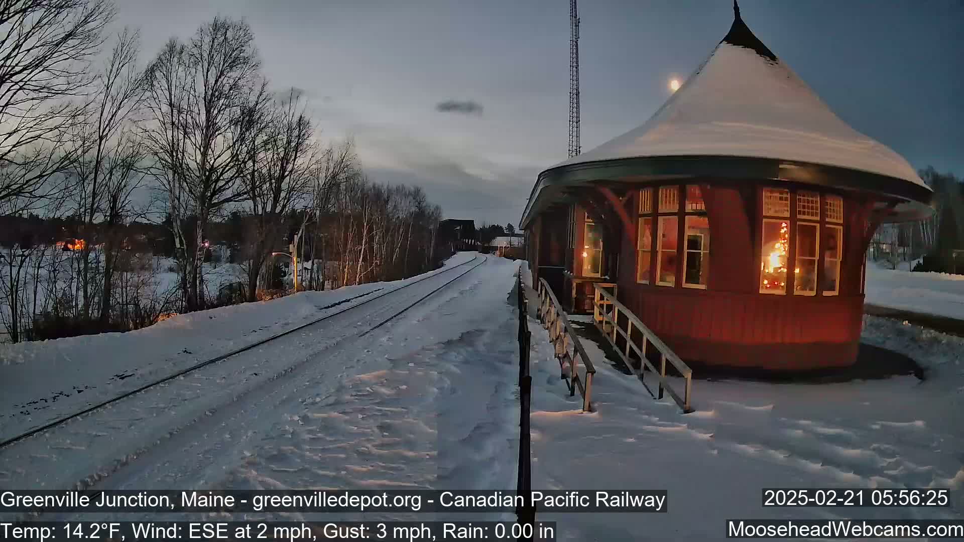 A snow-covered train platform with a red octagonal building, illuminated from within, sits beside train tracks under a twilight sky.