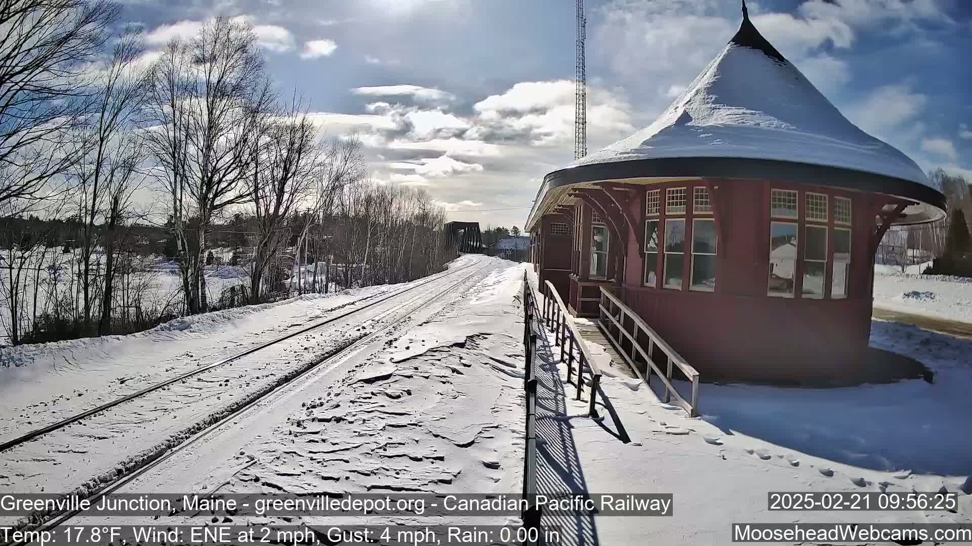 A snow-covered train station and railway track are seen on a partly sunny day with a light wind.