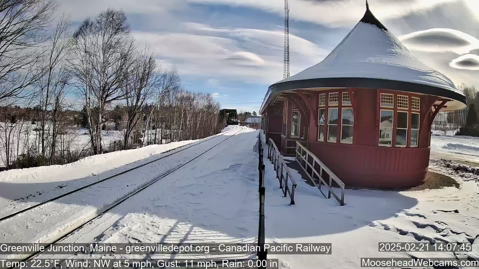 A snow-covered railway track leads to a red, round train station under a partly sunny sky with some lenticular clouds.
