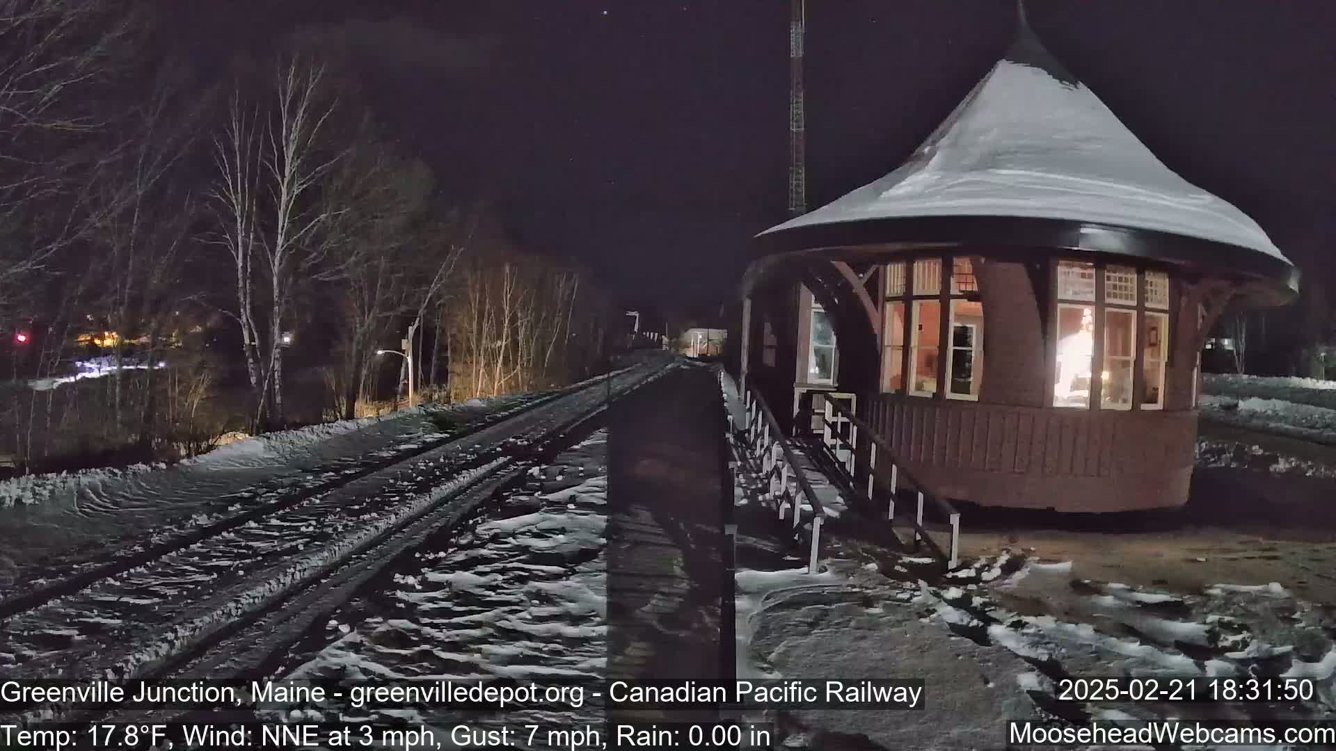 A snow-covered train platform and a lighted, snow-dusted, octagonal building are situated beside a snow-covered railway track at night.