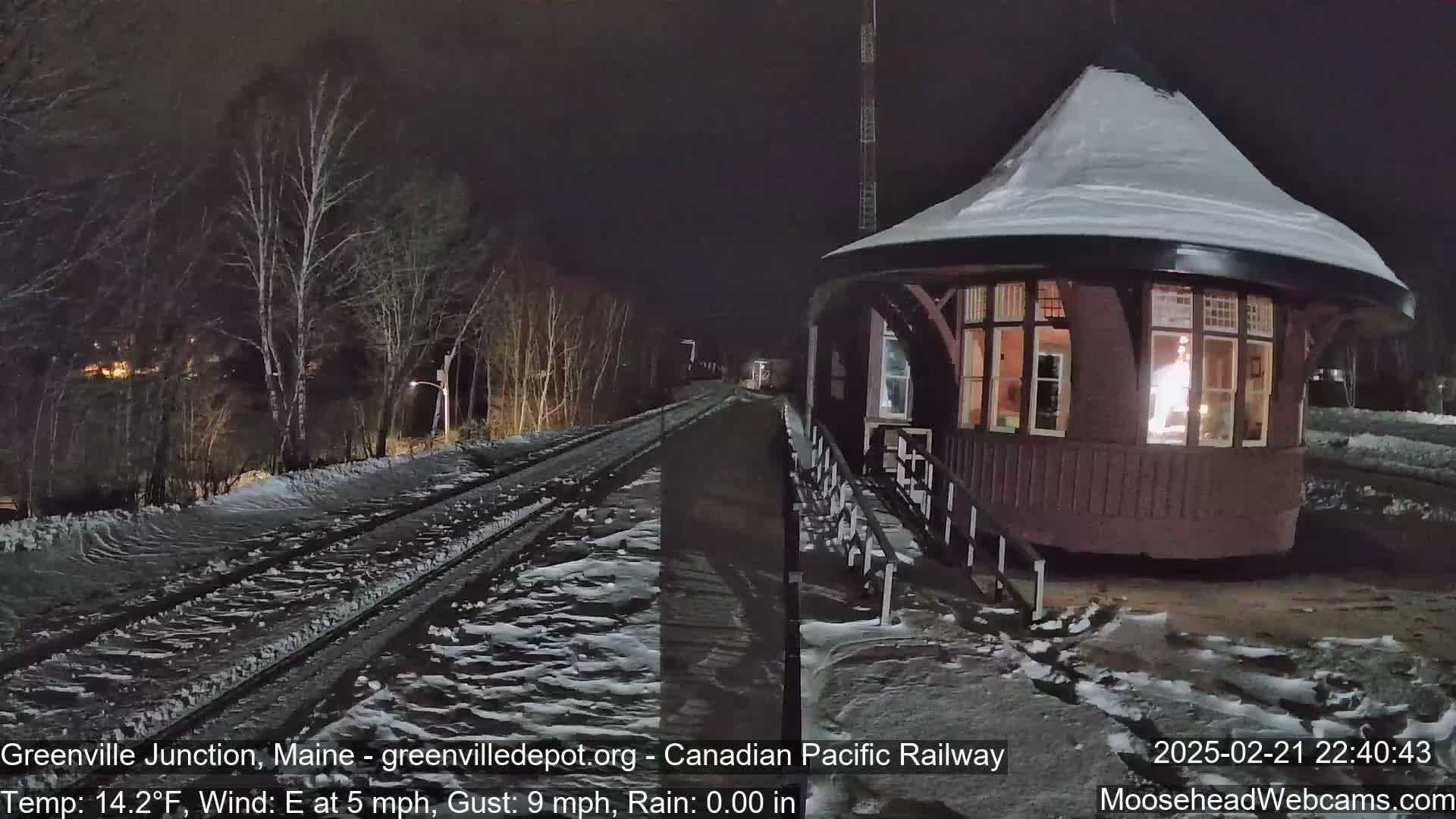 A snow-covered train platform and tracks are illuminated at night next to a small, snow-dusted building with lit windows.