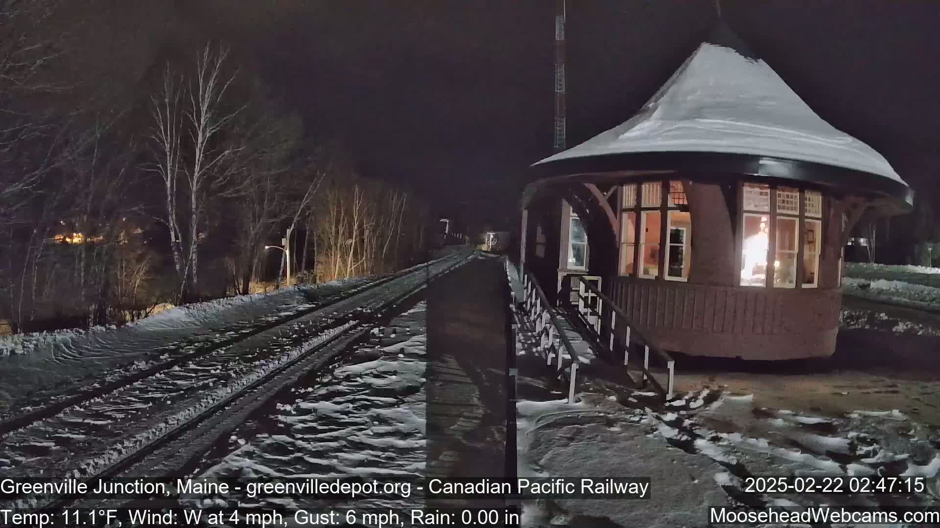 A snow-covered train platform with a round, lit building next to a set of train tracks at night.