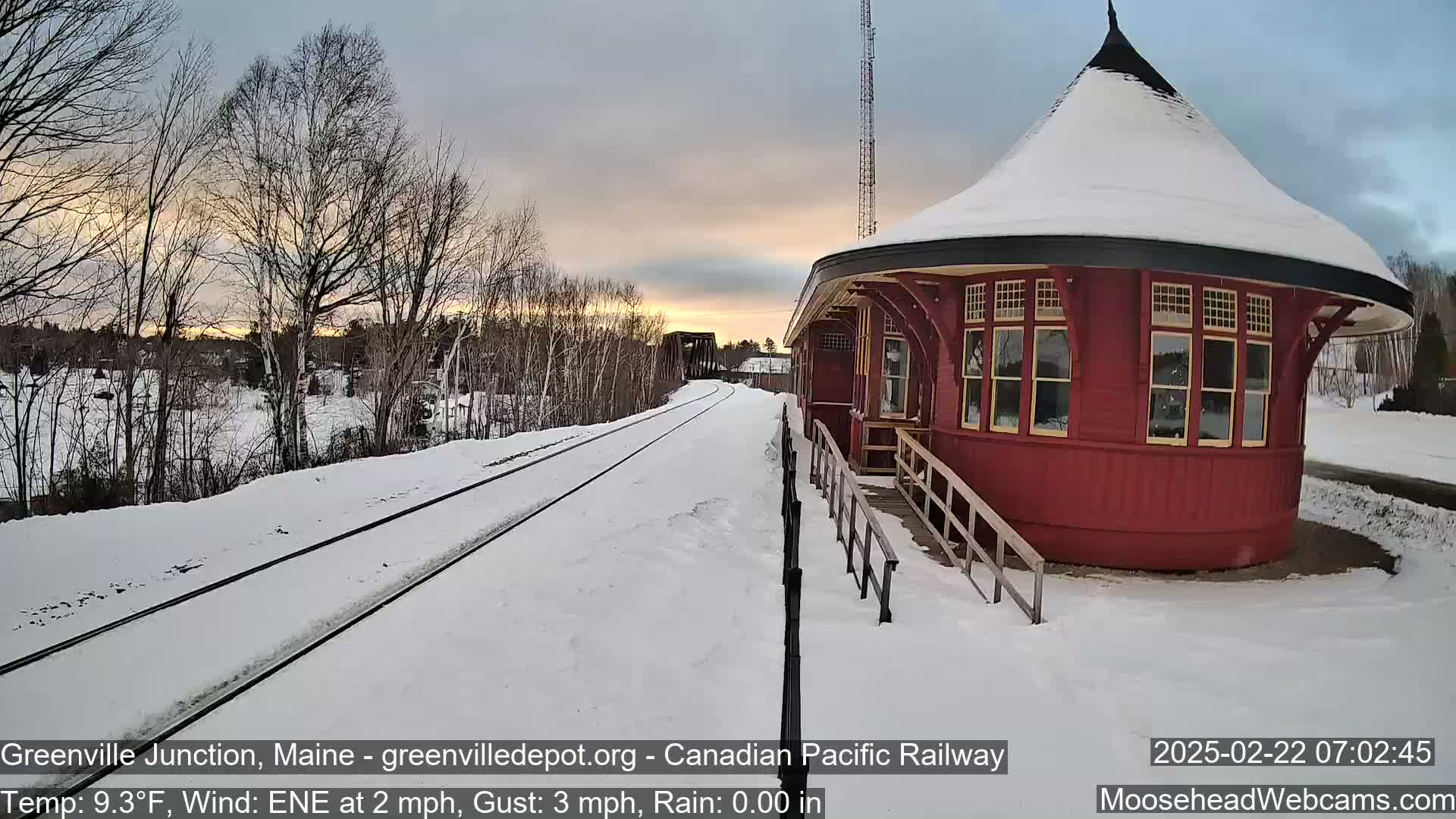 A red, snow-covered train depot sits beside snow-covered train tracks under a twilight sky.
