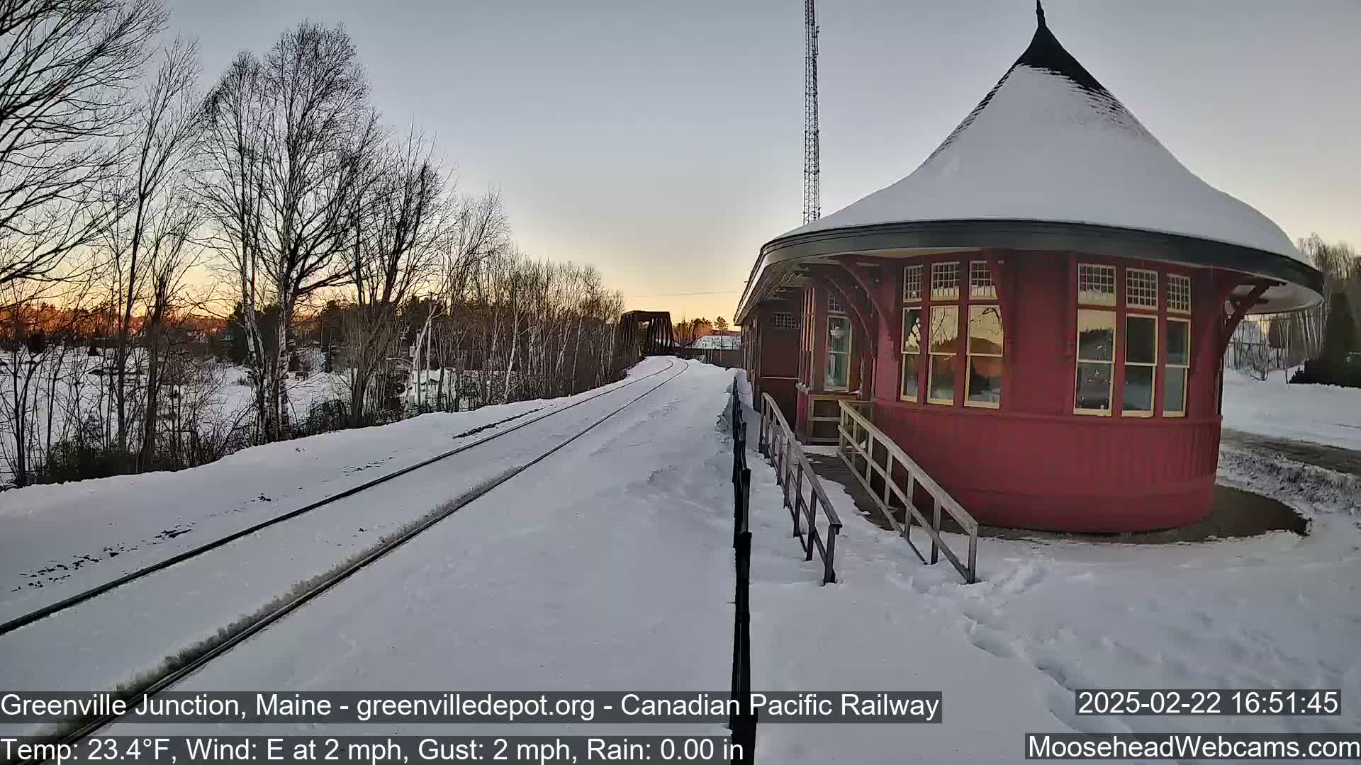 A snow-covered train station with a circular red waiting room sits beside a set of train tracks at sunset.