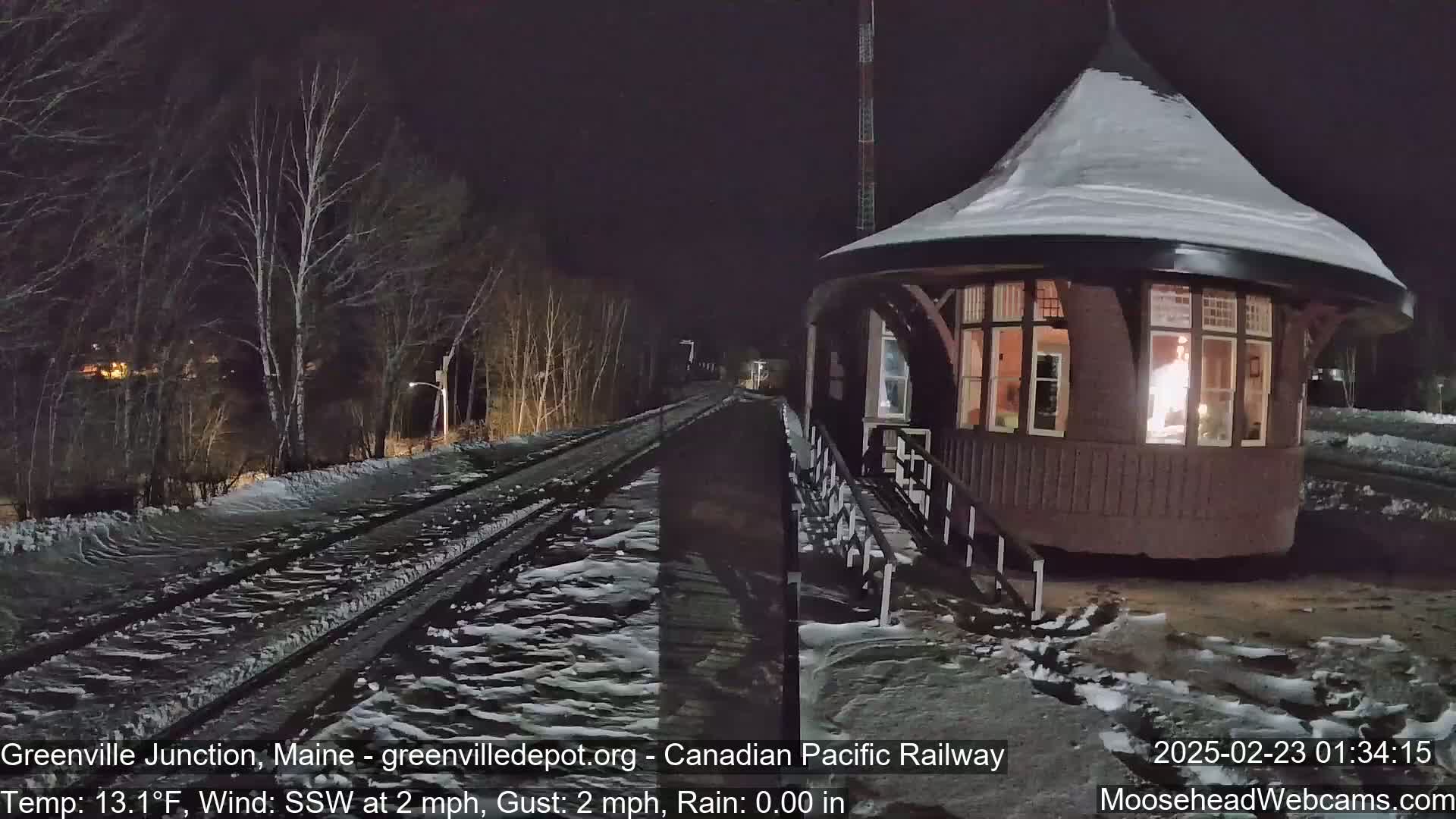 A snow-covered train station with a lit interior is situated beside a set of train tracks on a cold, windy night with light snow on the ground.