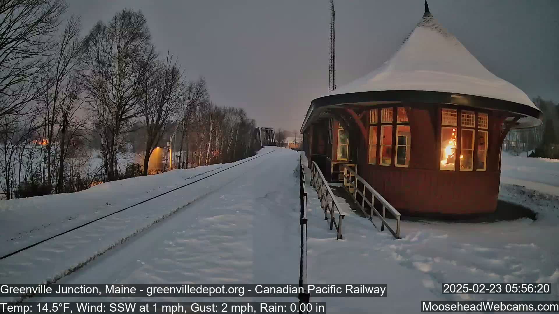 A snow-covered train station with a lit interior stands beside a snow-covered railway track under a twilight sky with a light south-southwesterly wind.