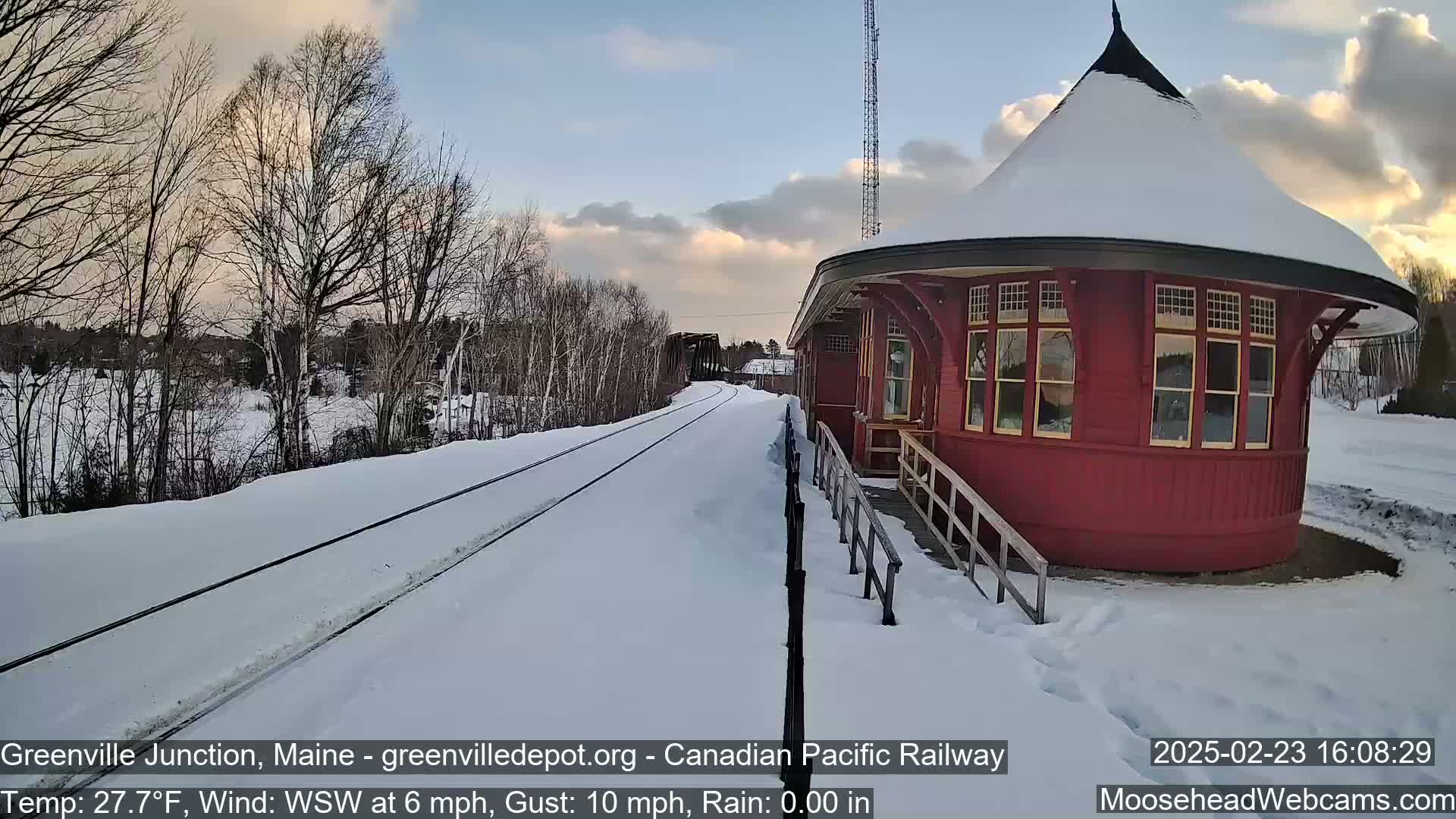 A snow-covered railway track leads to a red, round train station under a partly cloudy sky.