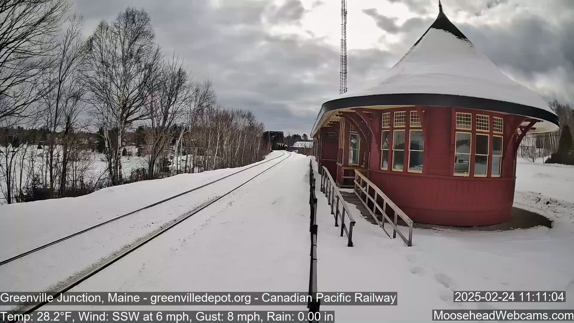Snow-covered railroad tracks extend toward a red, round train station under a cloudy sky.