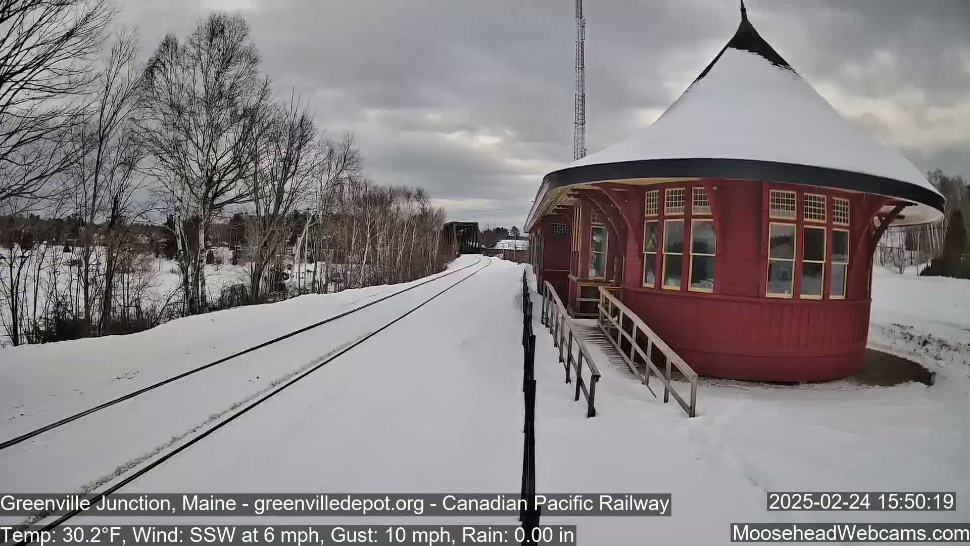 A snow-covered railroad track leads to a red, round train station under a cloudy sky.