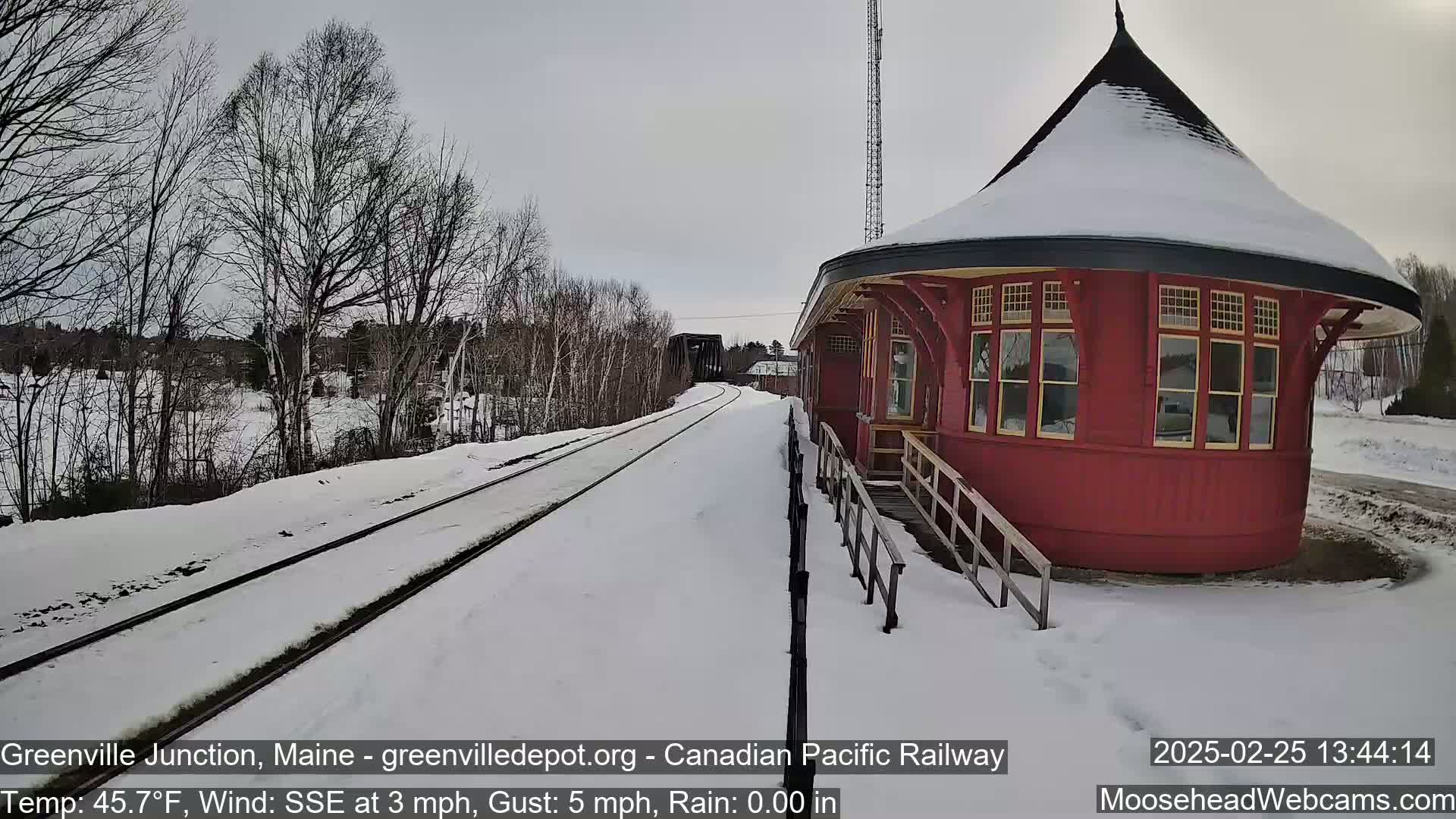 Snow-covered railroad tracks lead to a red, snow-topped train station in a winter scene with a light wind.