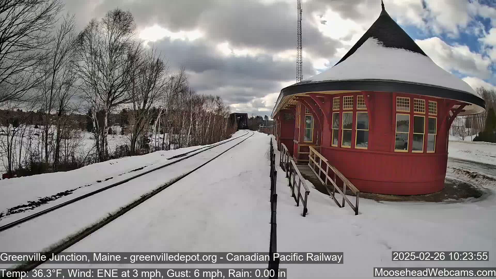 A snow-covered train platform with a red, round train station building is visible under a cloudy sky.