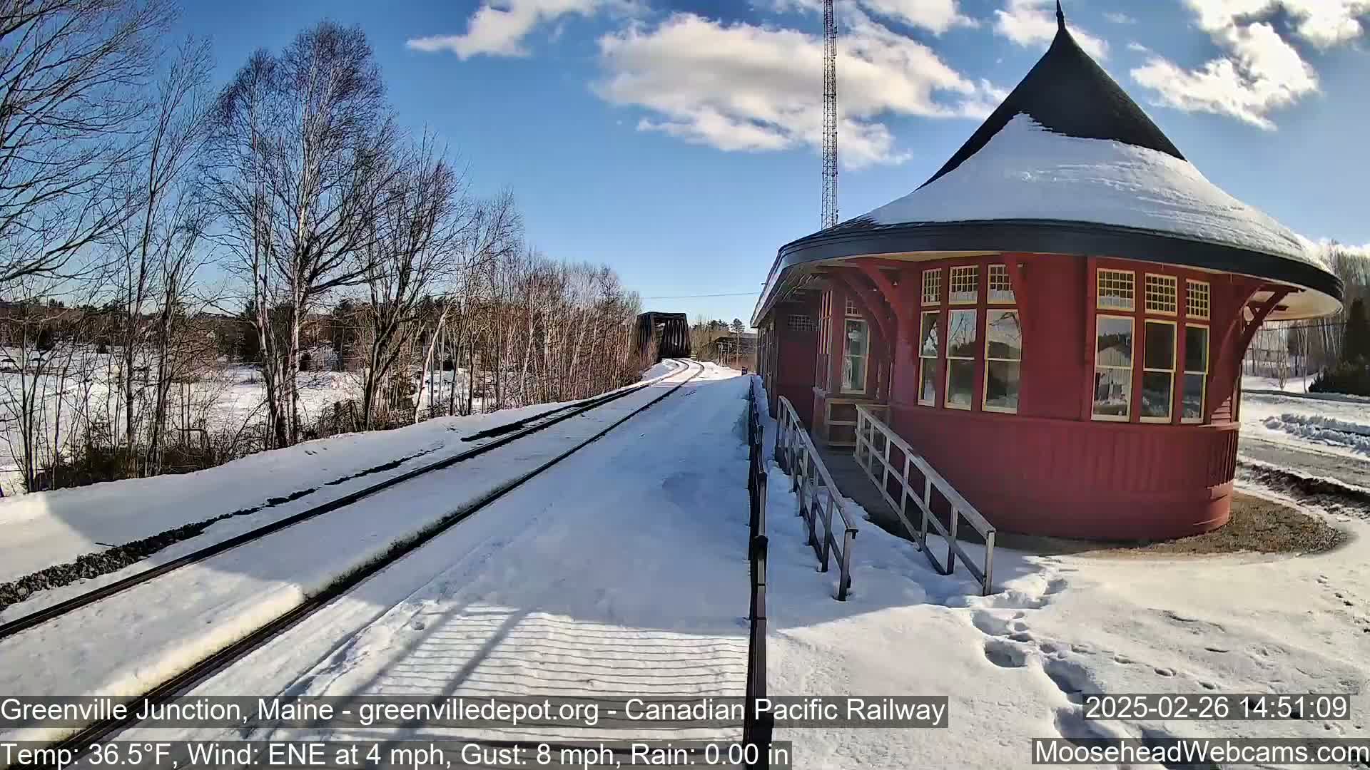 A red, octagonal train depot sits beside snow-covered train tracks on a partly sunny day with a light wind.