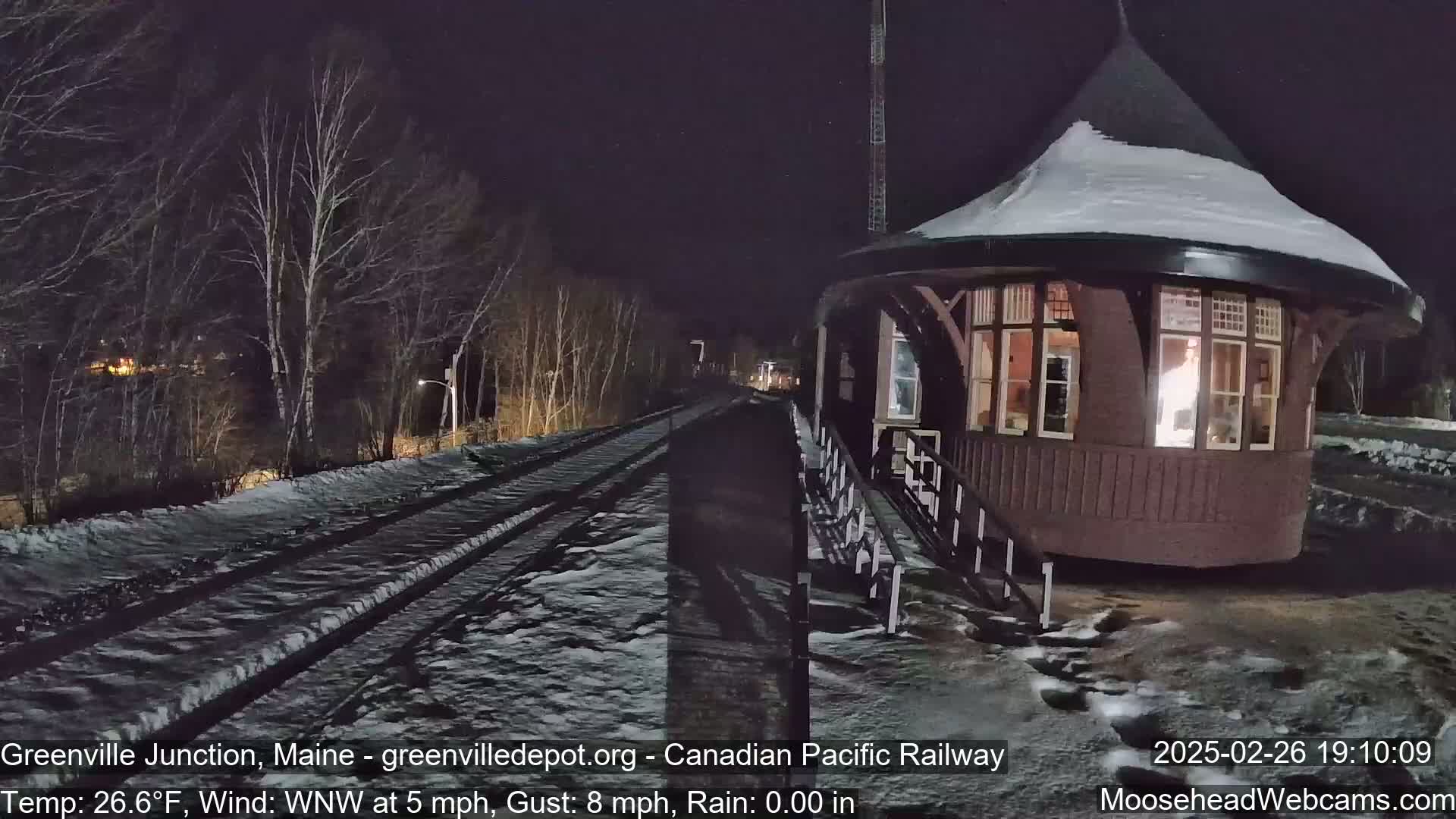 A snow-covered train platform and a small, illuminated building are visible at night, with a light wind blowing.
