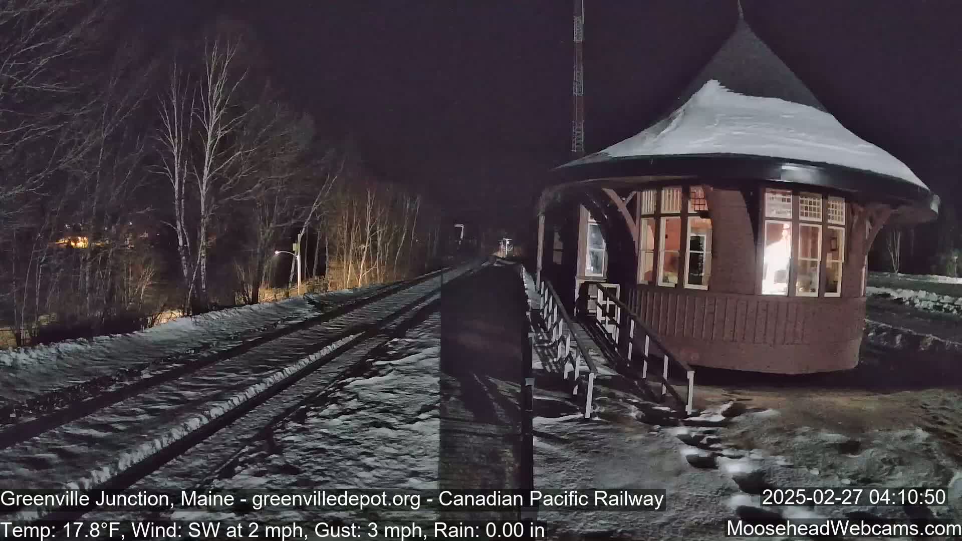 A snow-covered train platform and tracks are visible at night, next to a small, illuminated, octagonal building under a dark sky with a light southwest wind.