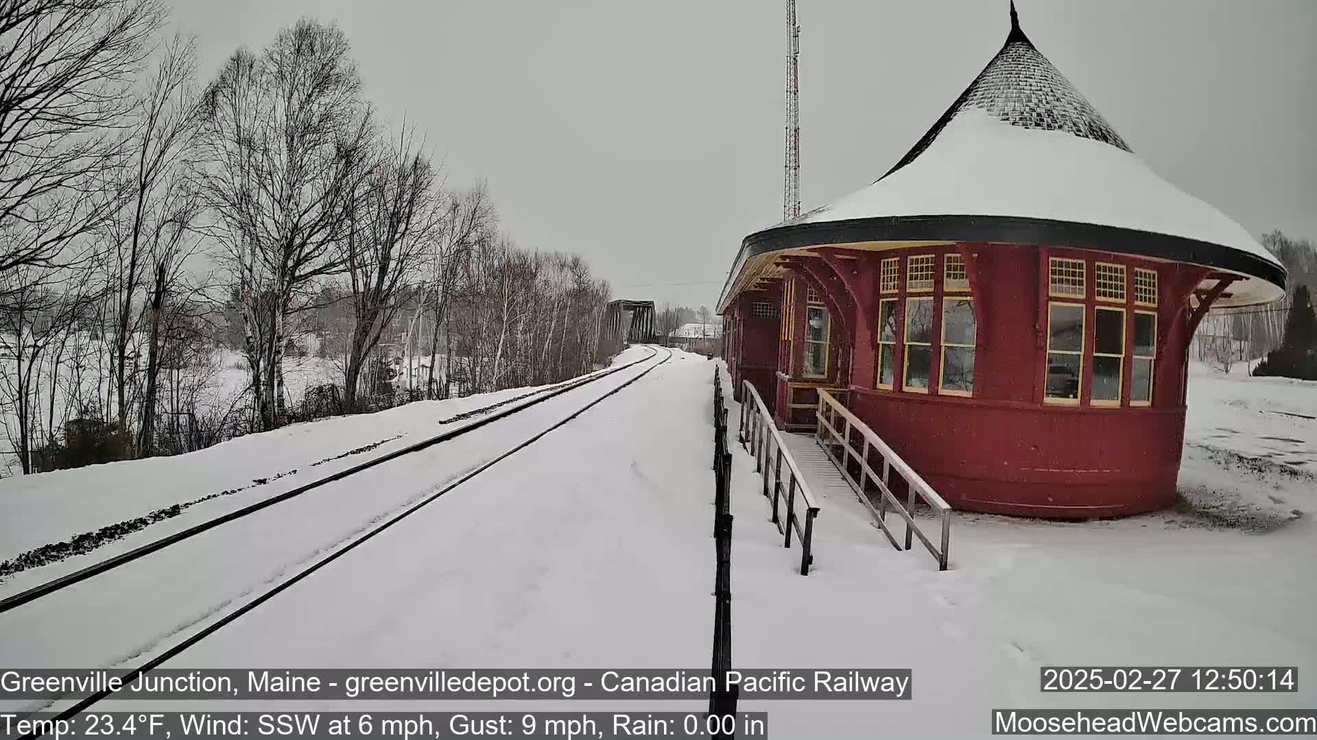 Snow-covered railroad tracks lead to a red, snow-dusted train depot in a snowy landscape.