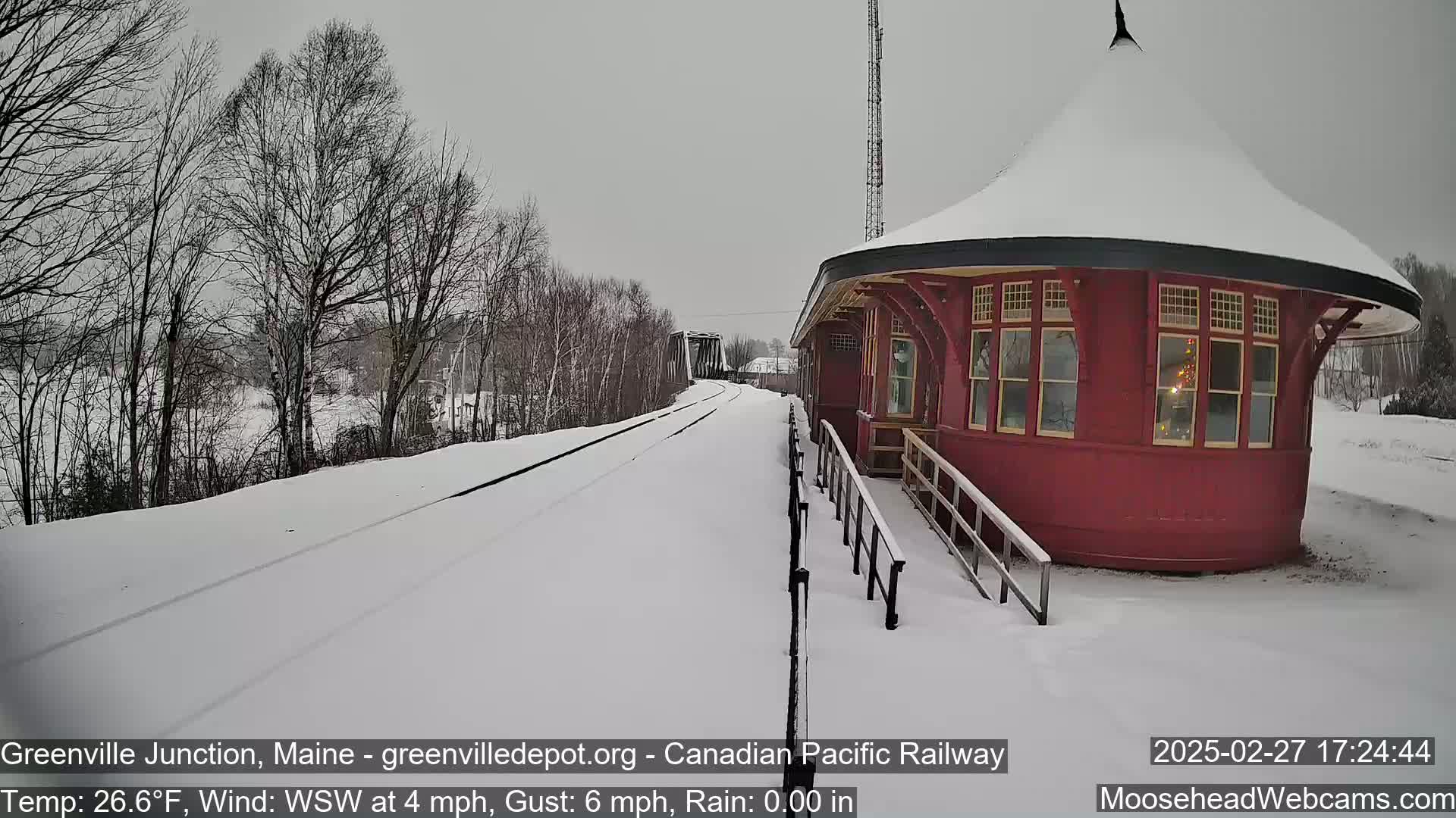 A snow-covered railway platform with a red octagonal building and bare trees under an overcast sky.