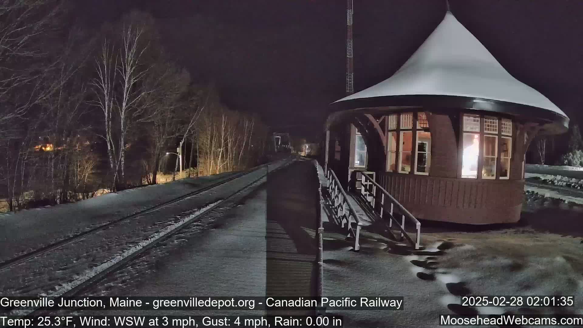 A snow-covered train platform and tracks are illuminated at night under a dark sky, with bare trees lining the platform.
